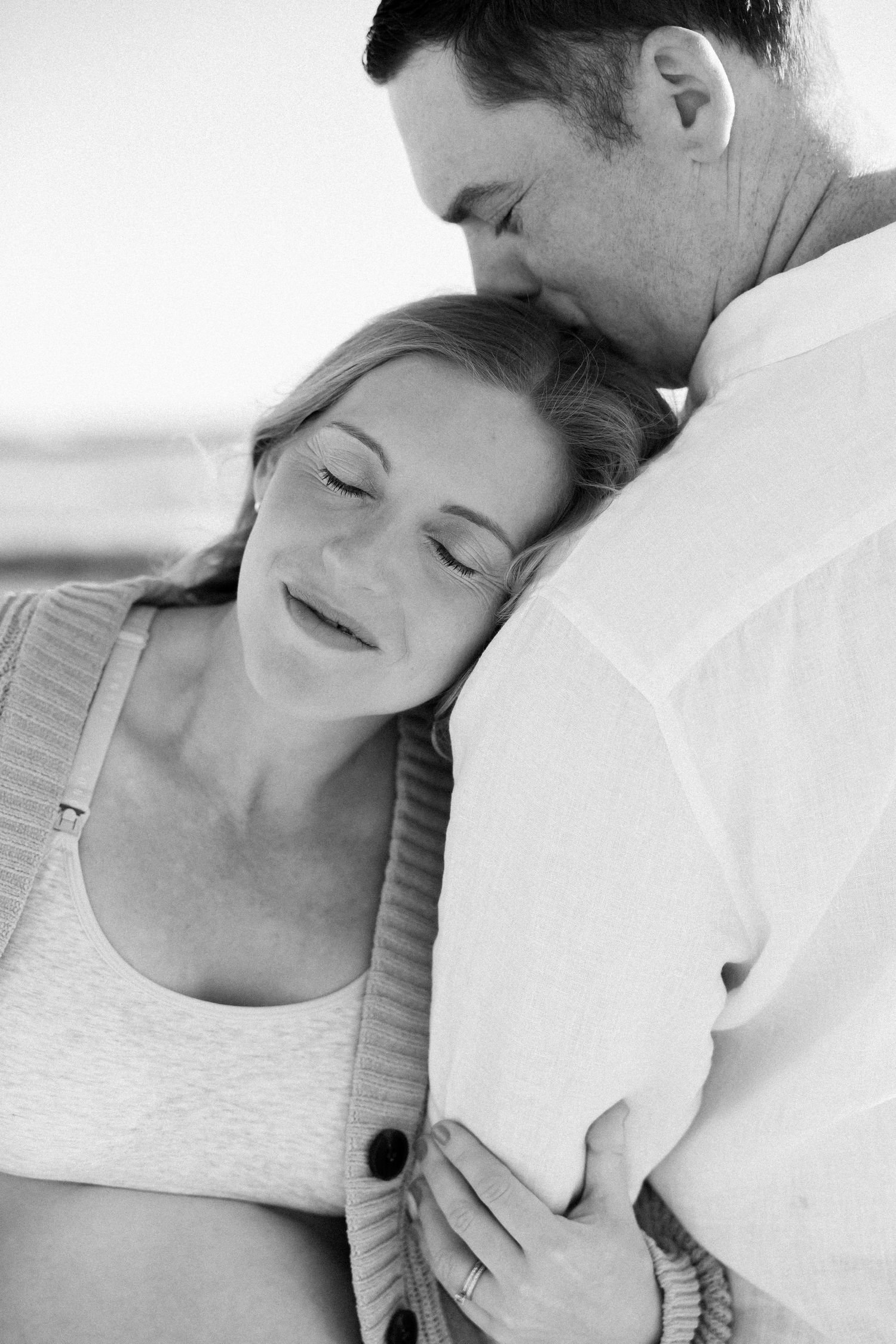 Black and white photo of man kissing pregnant woman on forehead as she smiles with eyes closed.