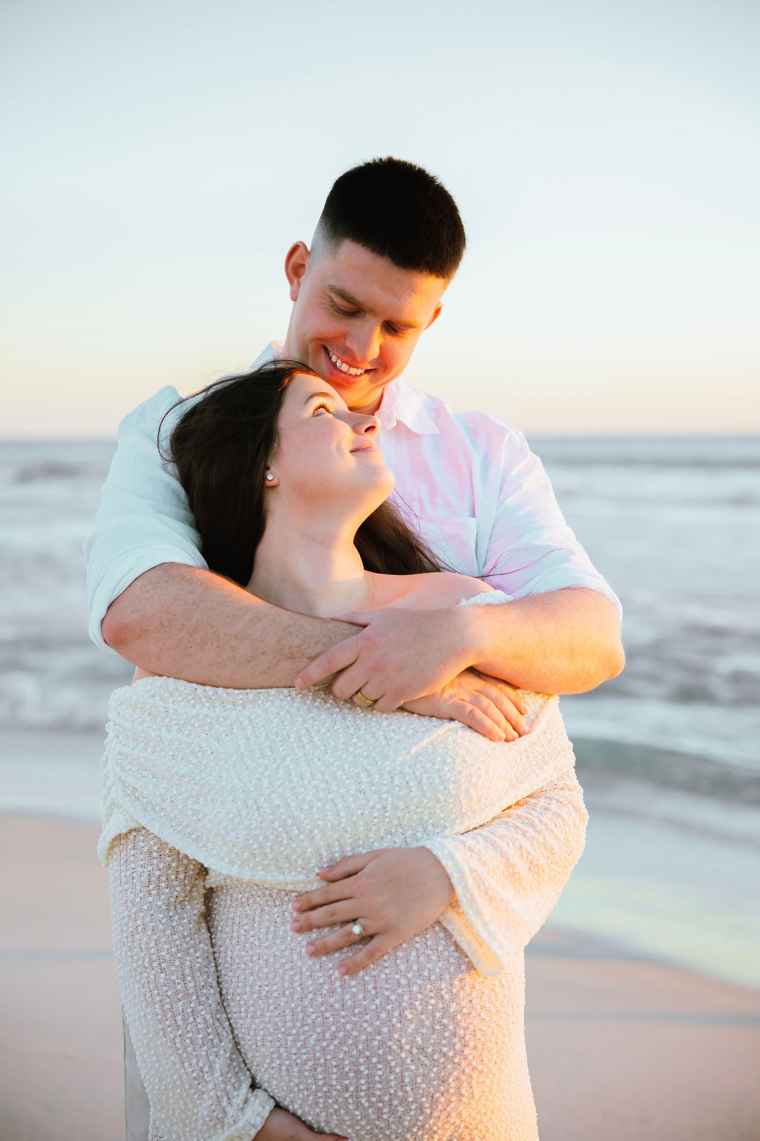 Man in white shirt embracing pregnant woman from behind on beach, both smiling at golden hour.