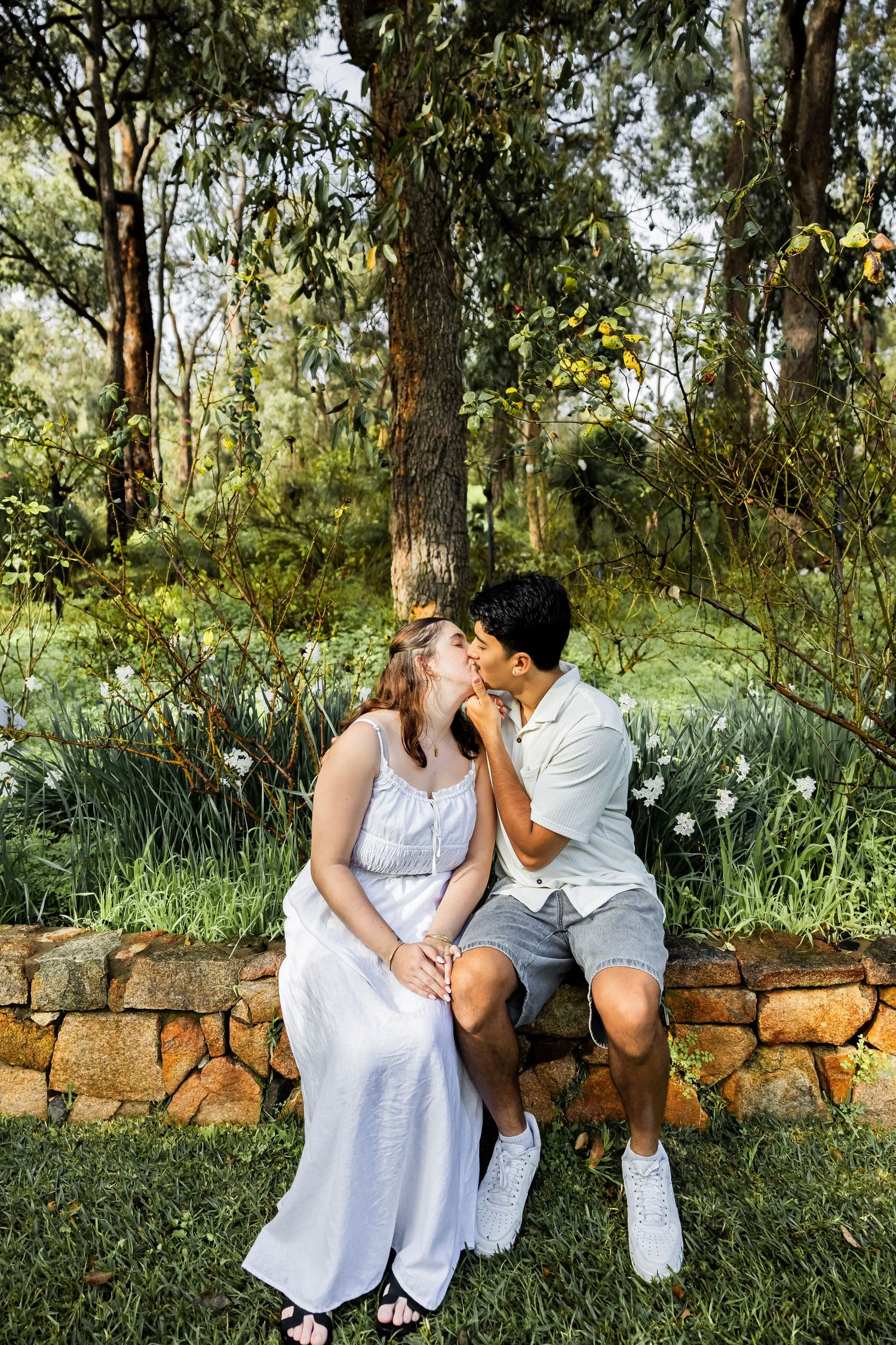 Couple sitting on a stone wall sharing a kiss in a lush green garden setting.