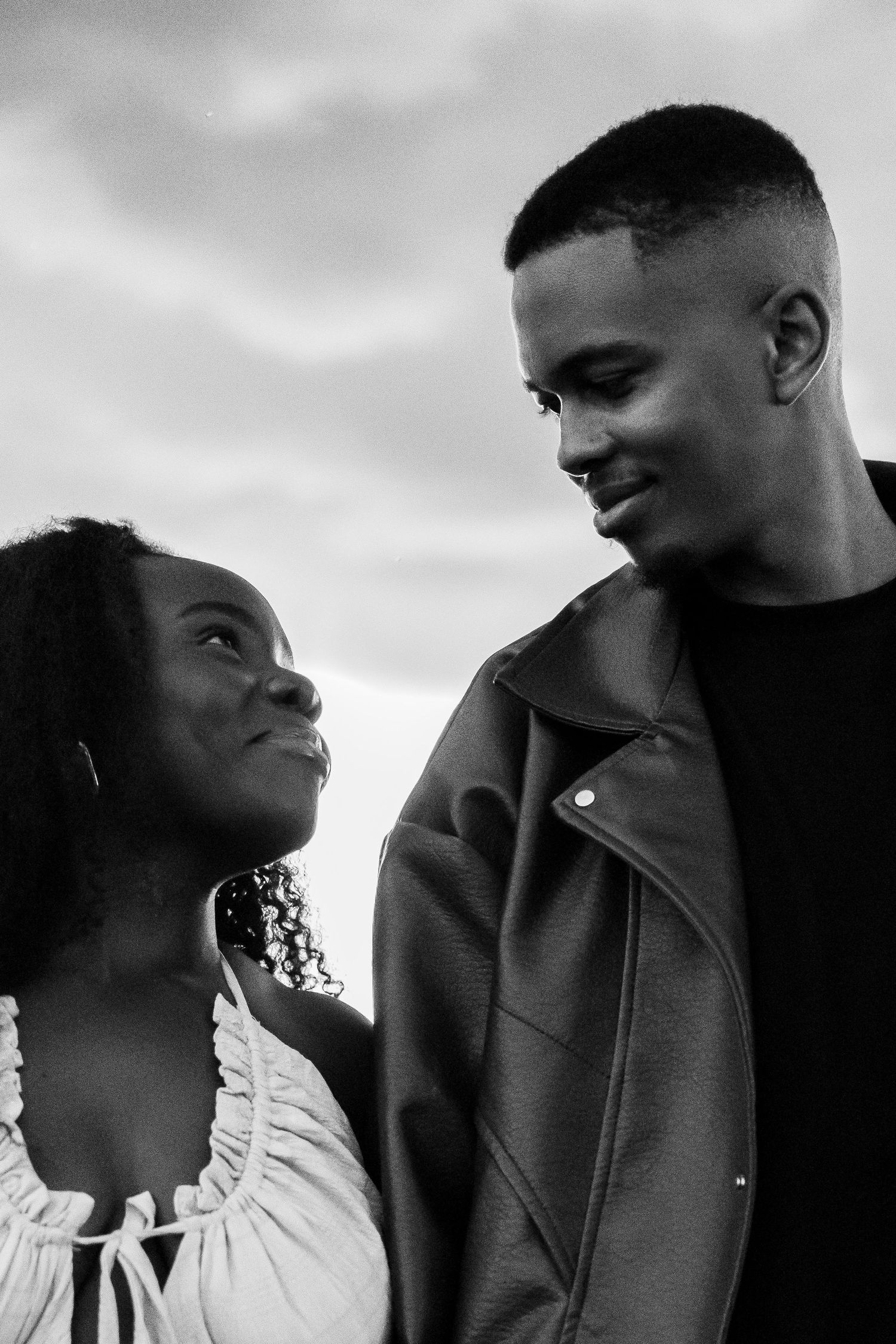Black and white portrait of a smiling young couple gazing at each other outdoors under a cloudy sky.