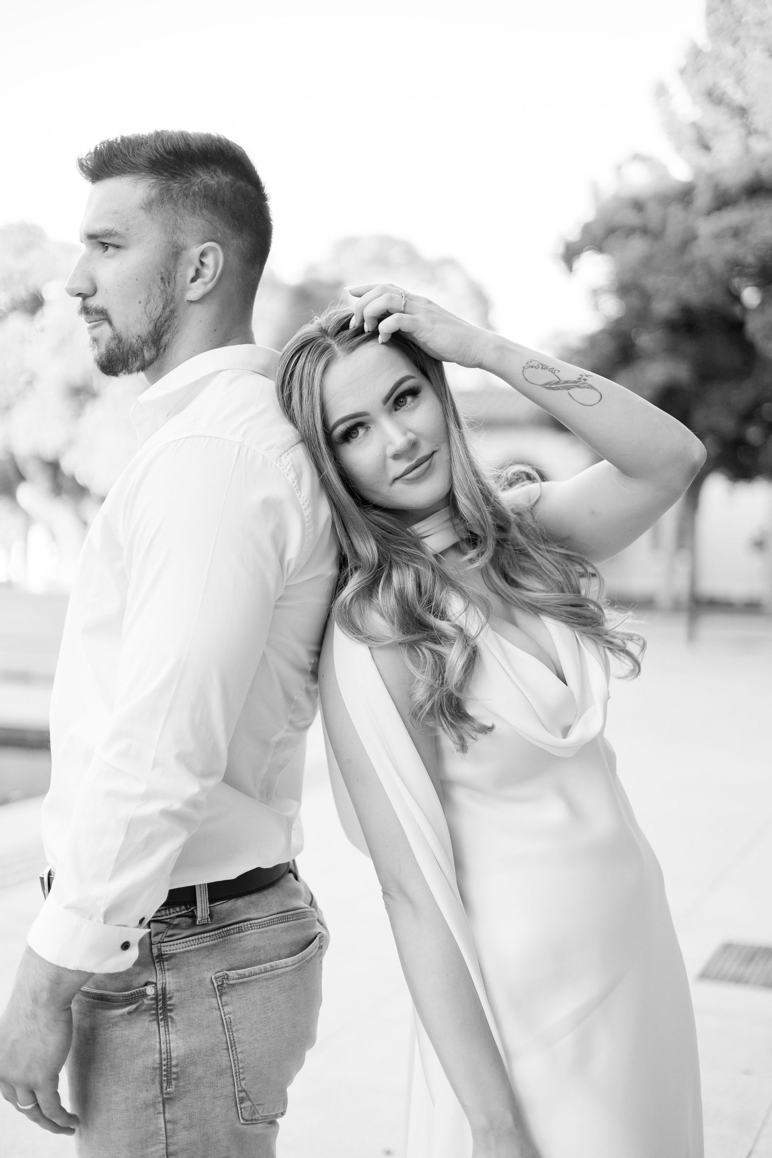 Black and white portrait of couple back-to-back, woman posing with hand in hair wearing white dress.