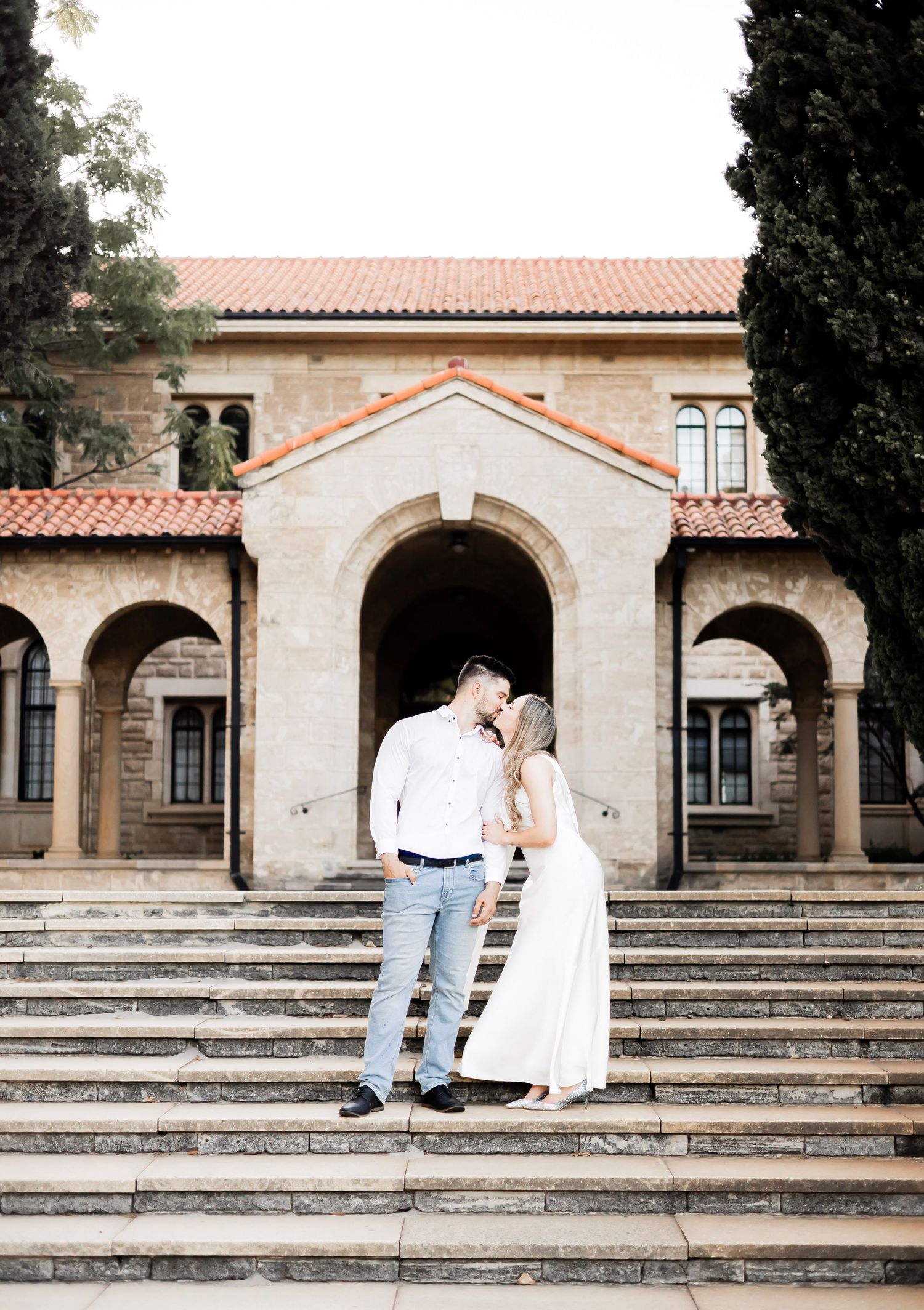 Couple sharing a kiss on grand stone steps in front of a historic Spanish Colonial Revival building.