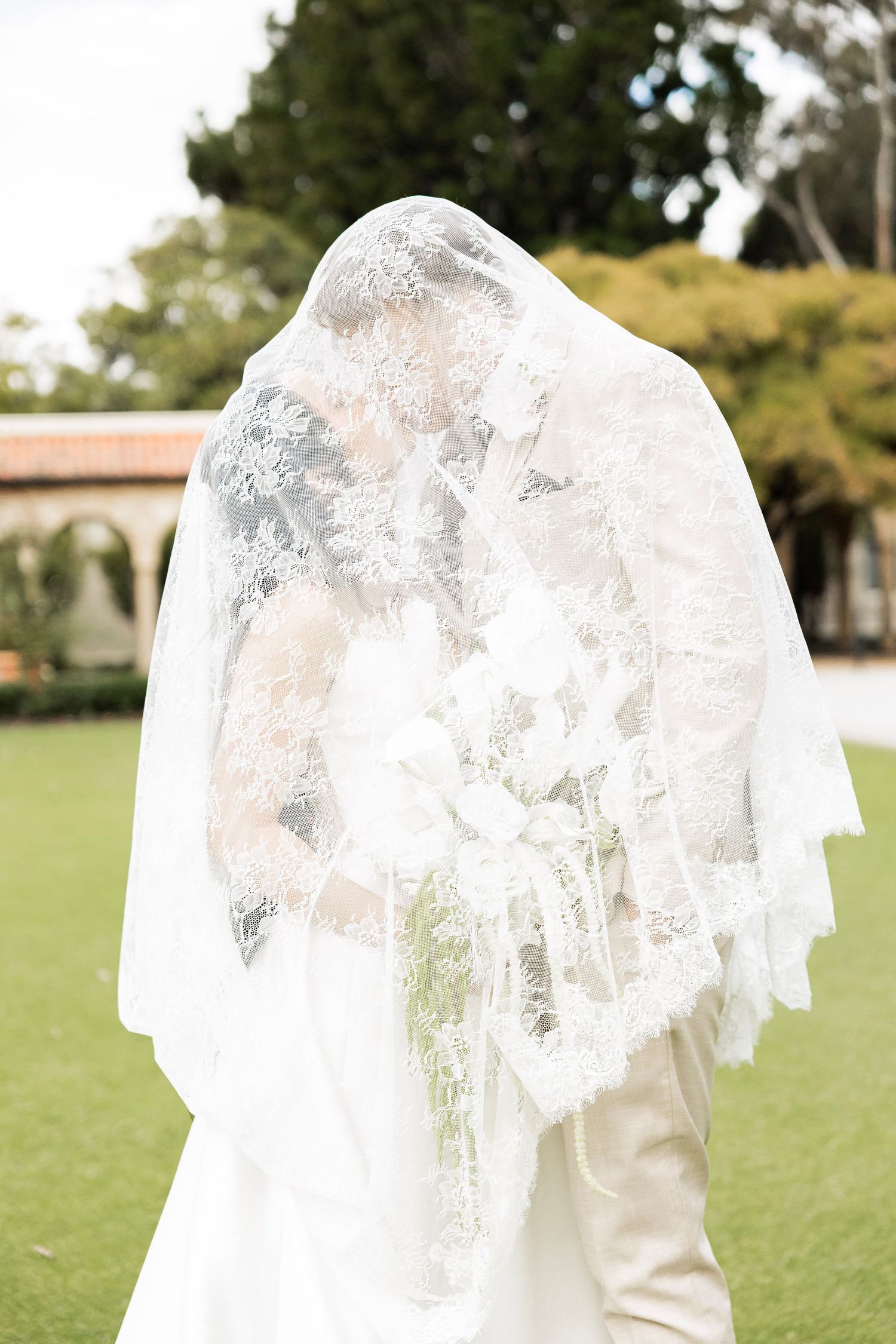 Bride and groom kiss under flowing lace veil outdoors at garden wedding venue.