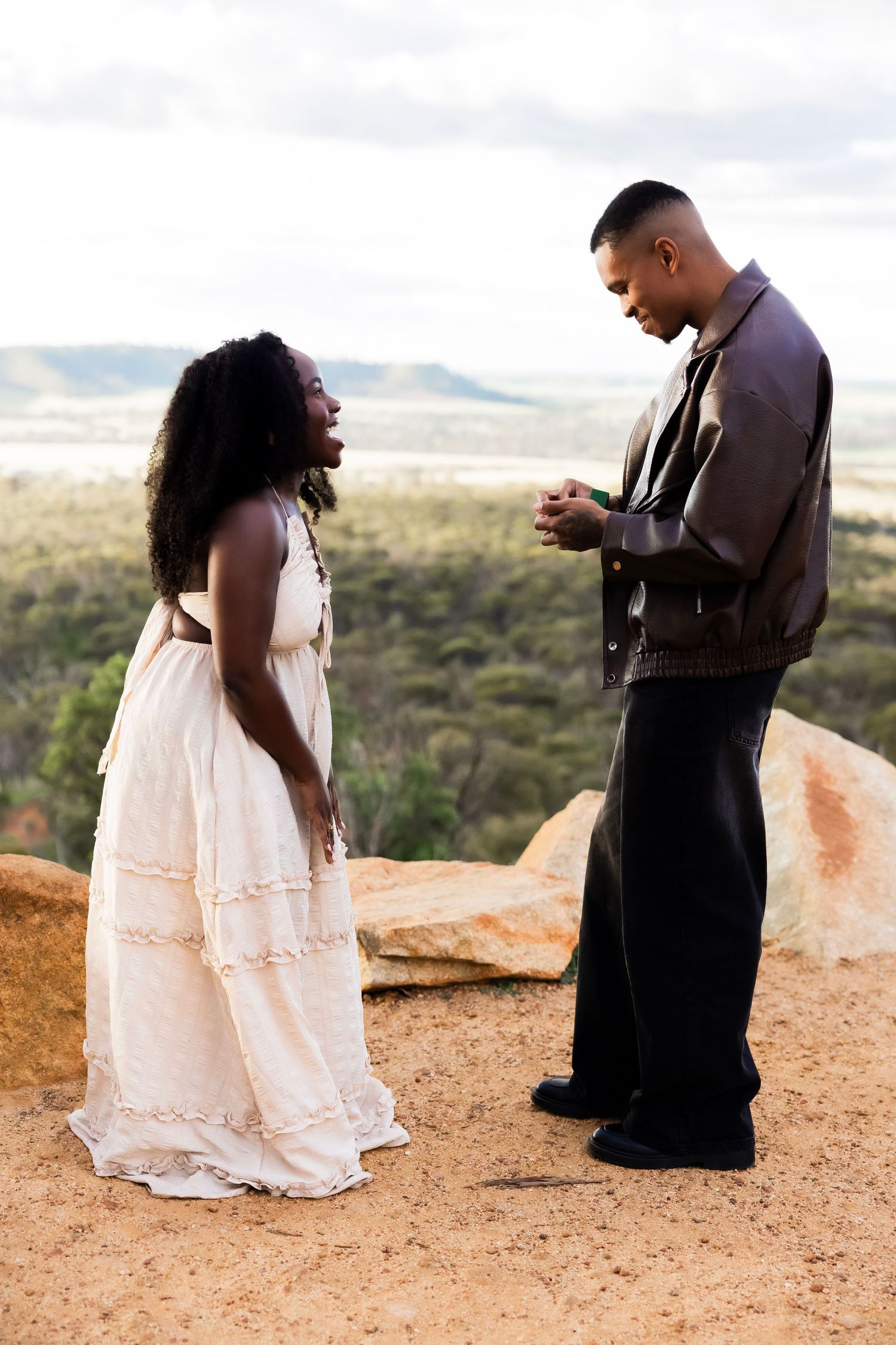 A couple stands on a rocky hilltop, woman in white dress smiling as man holds her hands during a romantic outdoor proposal.