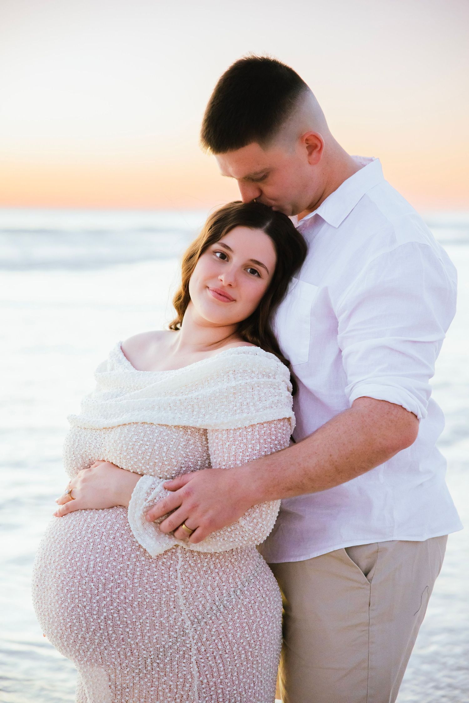 Man tenderly embracing pregnant woman from behind at golden hour beach maternity photoshoot.