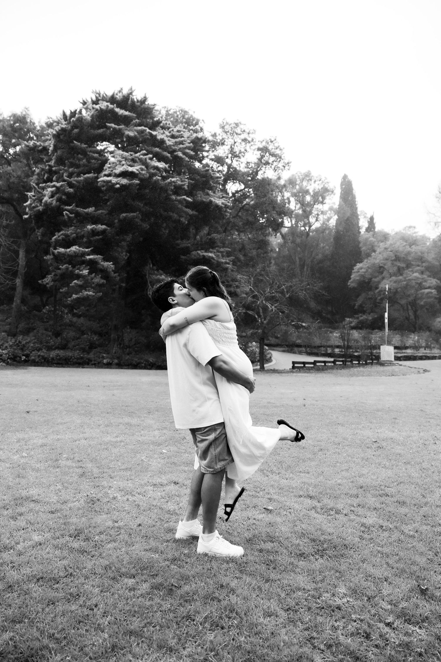 Black and white photo of a man lifting his partner in a grassy park surrounded by trees.