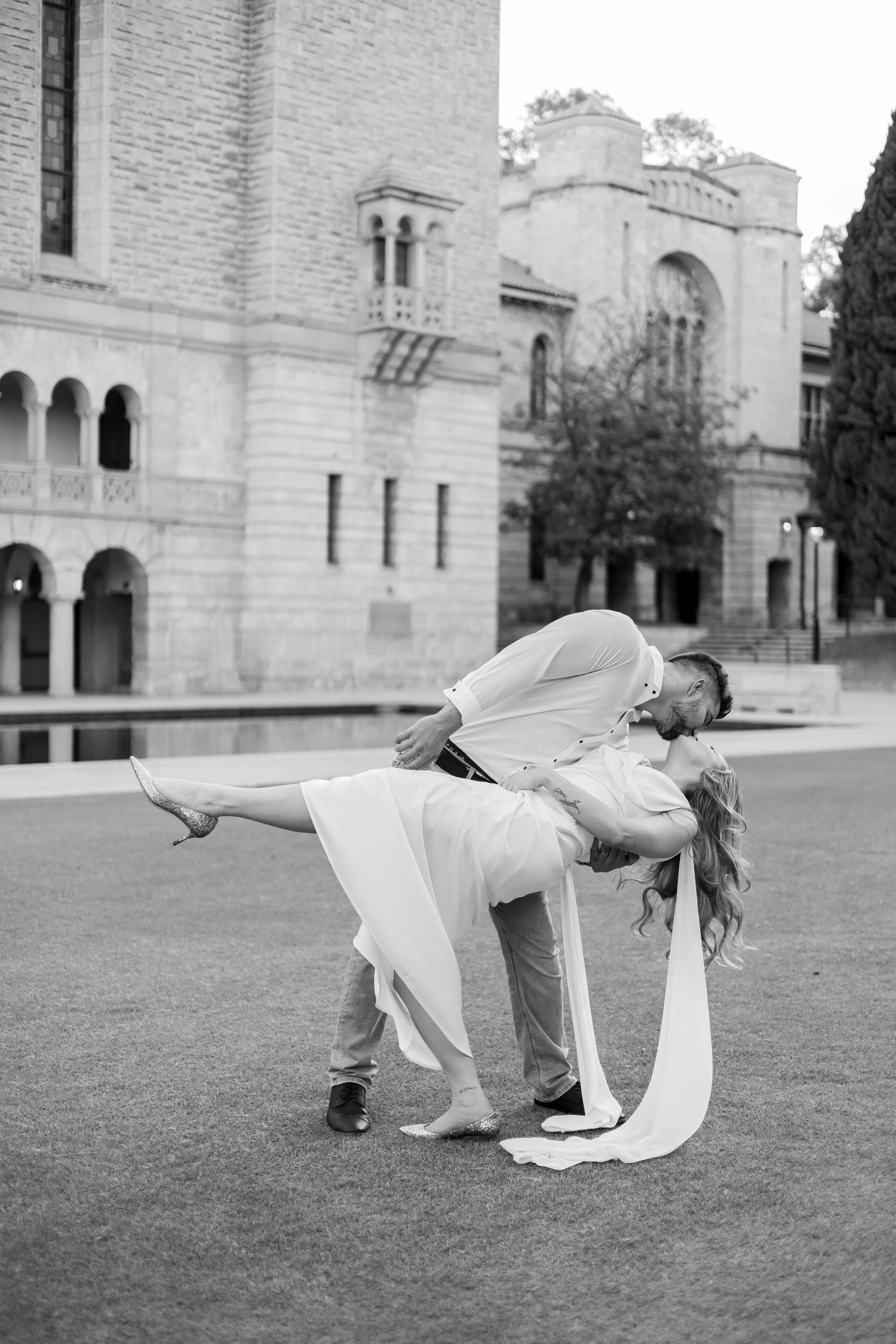 Black and white photo of groom dipping and kissing bride in white dress outdoors near historic stone building.