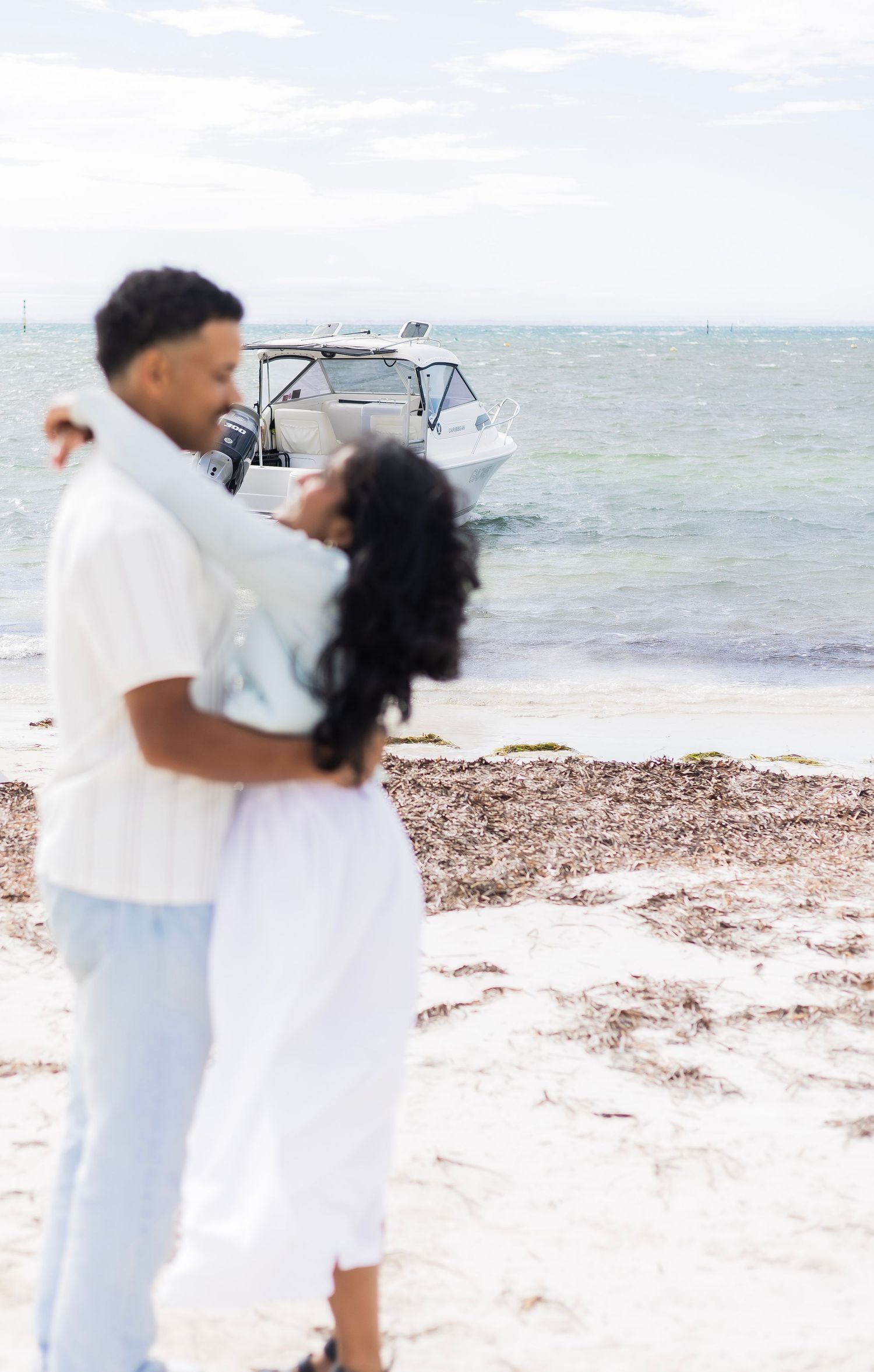 A couple embraces on a sandy beach with a motorboat floating on the ocean in the background.