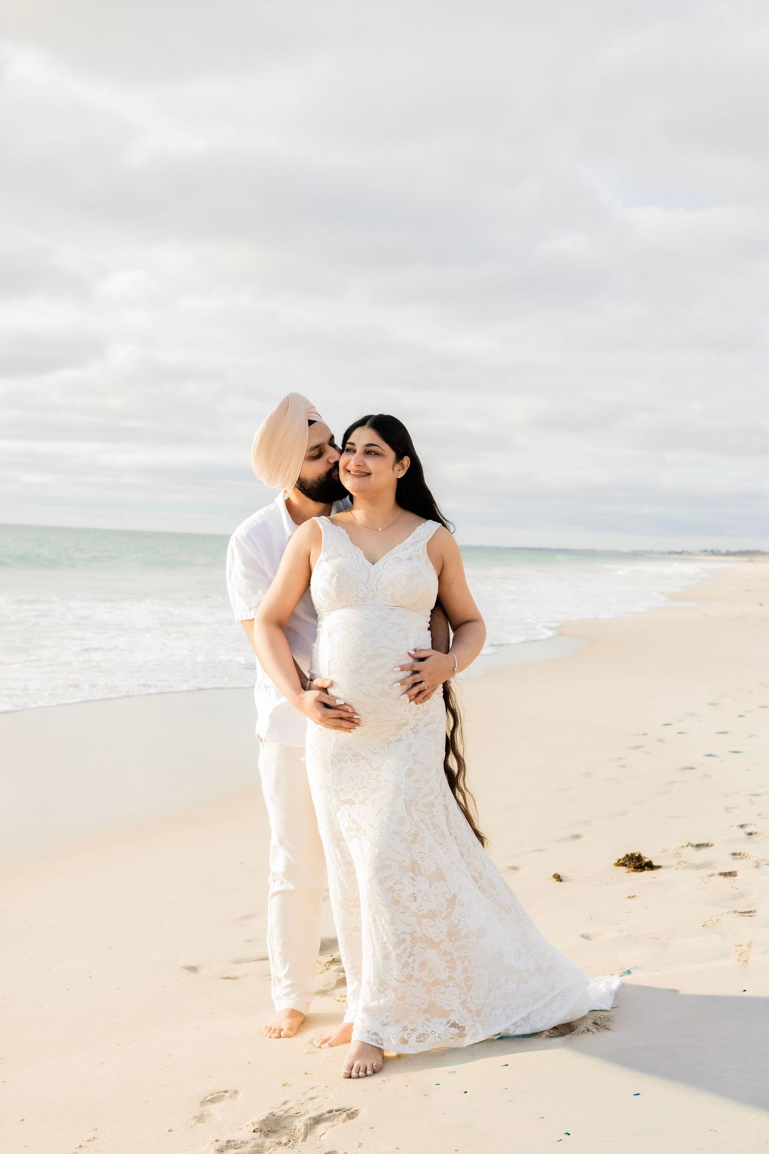 Expecting couple standing together on sandy beach, man in turban embracing pregnant partner in white lace gown.