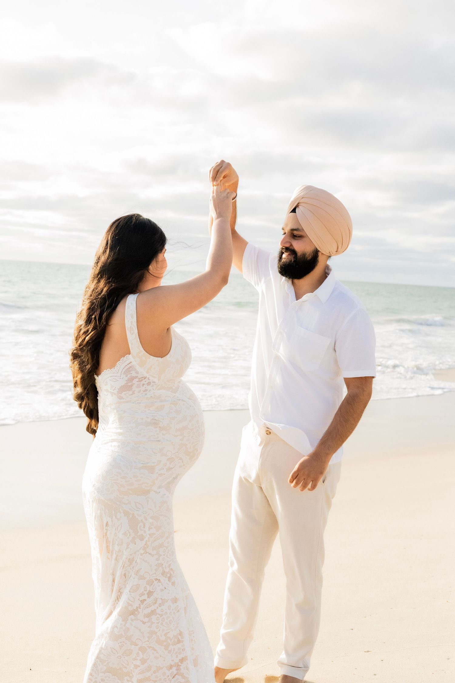 Expecting couple in white outfits dancing and holding hands on a bright sandy ocean beach.