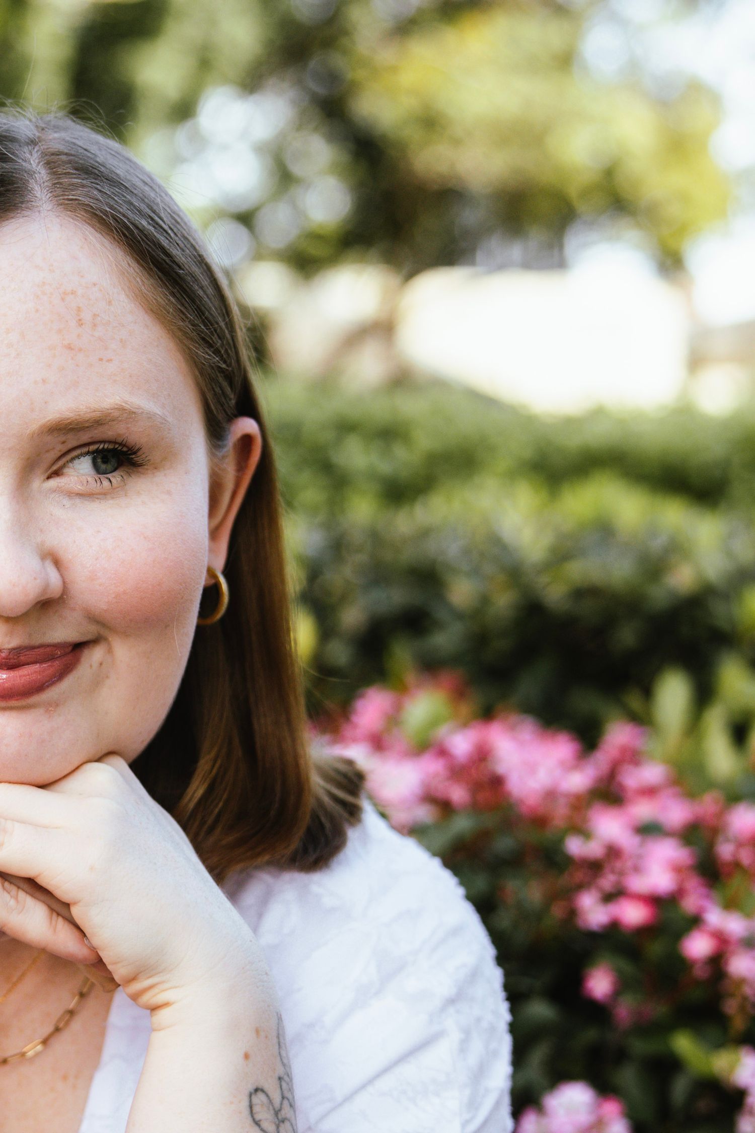 Young woman with freckles and gold hoop earrings resting chin on hands outdoors near pink flowers.