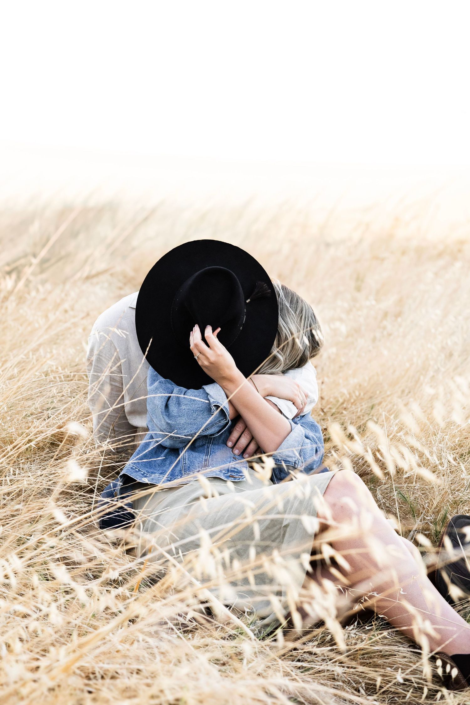 Couple sharing a hidden kiss behind a black cowboy hat while sitting in a grass field.