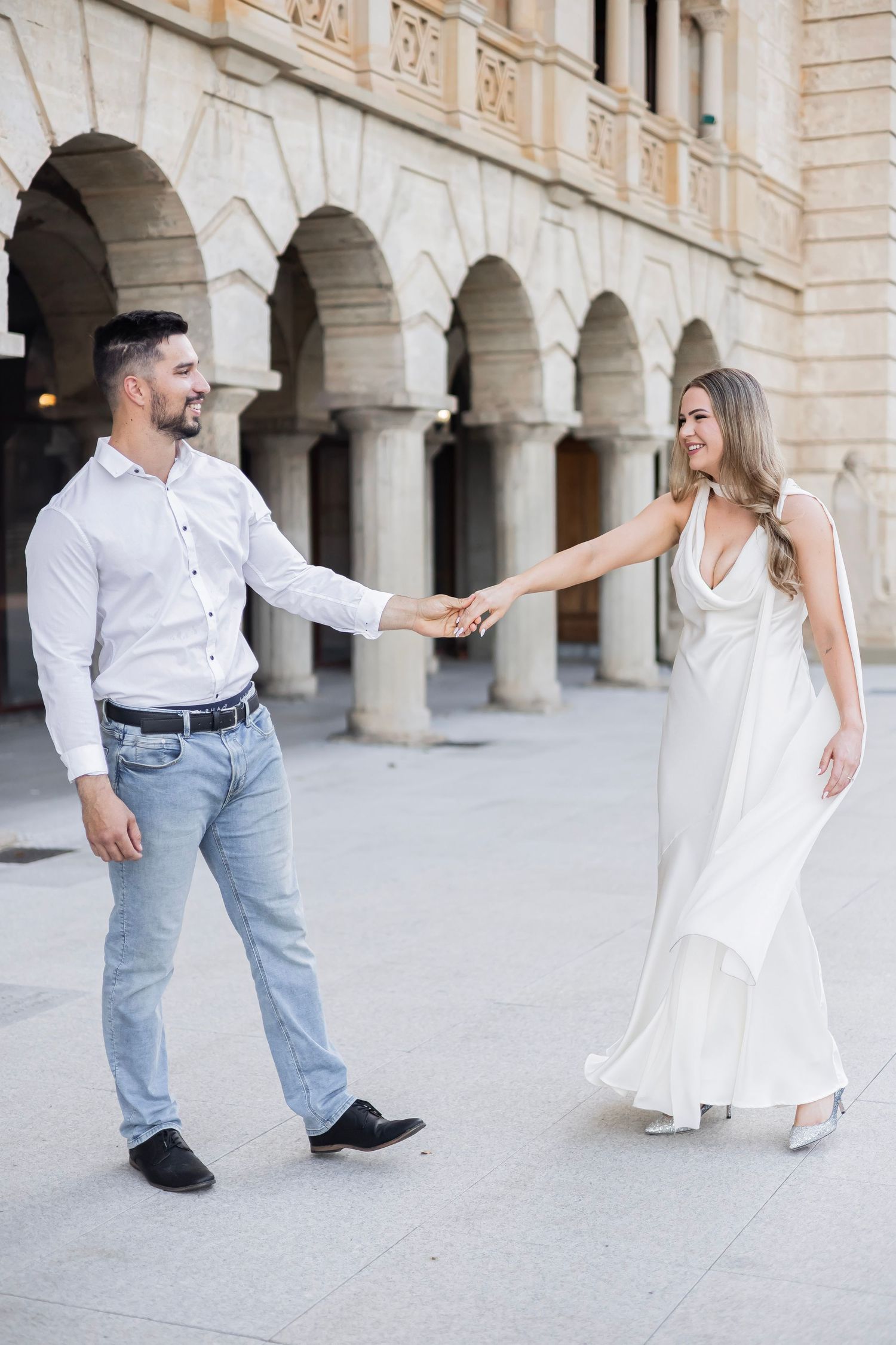 Happy couple holding hands and smiling at each other in front of elegant arched stone building.