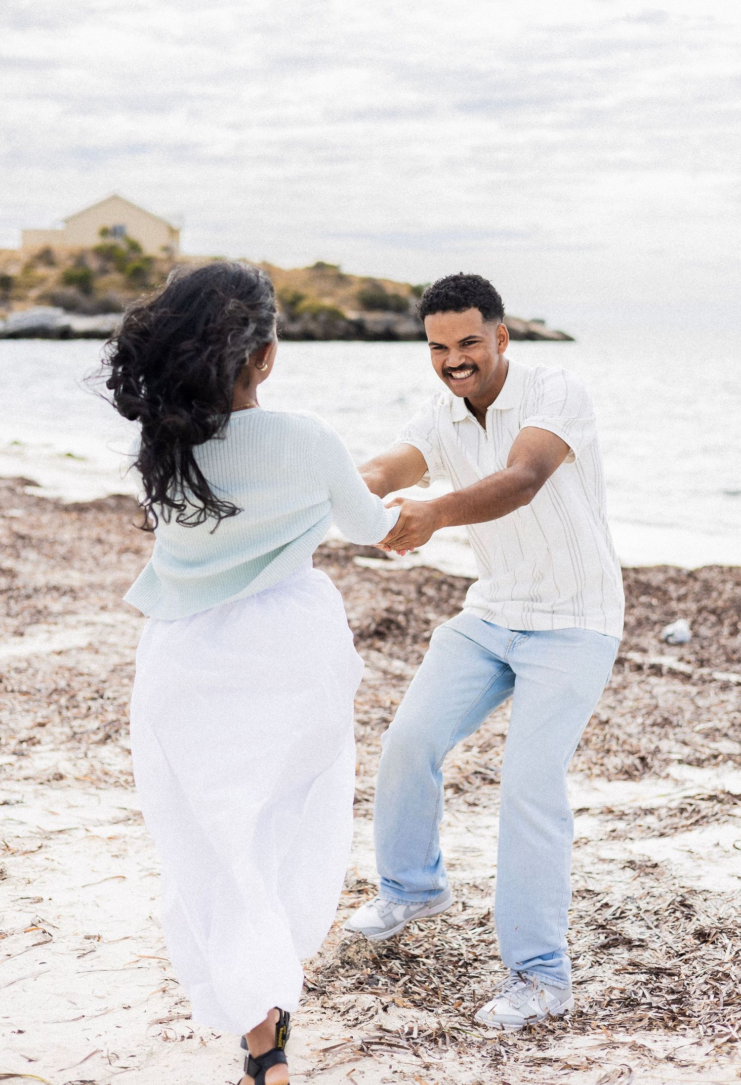 Couple holding hands and dancing playfully on a rocky beach with ocean waves behind them.