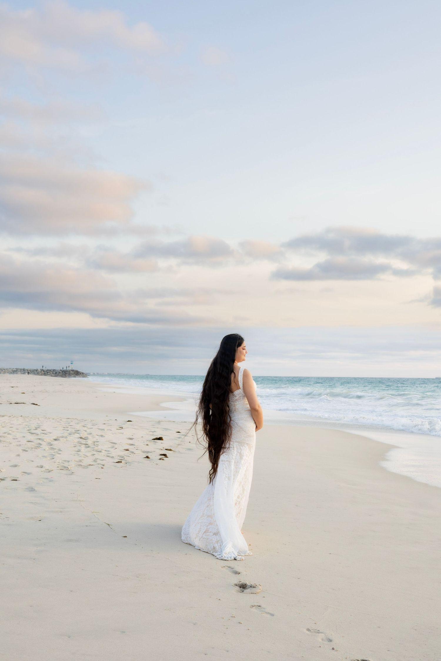 Pregnant woman in elegant white lace gown with long hair walking along beach at dusk.