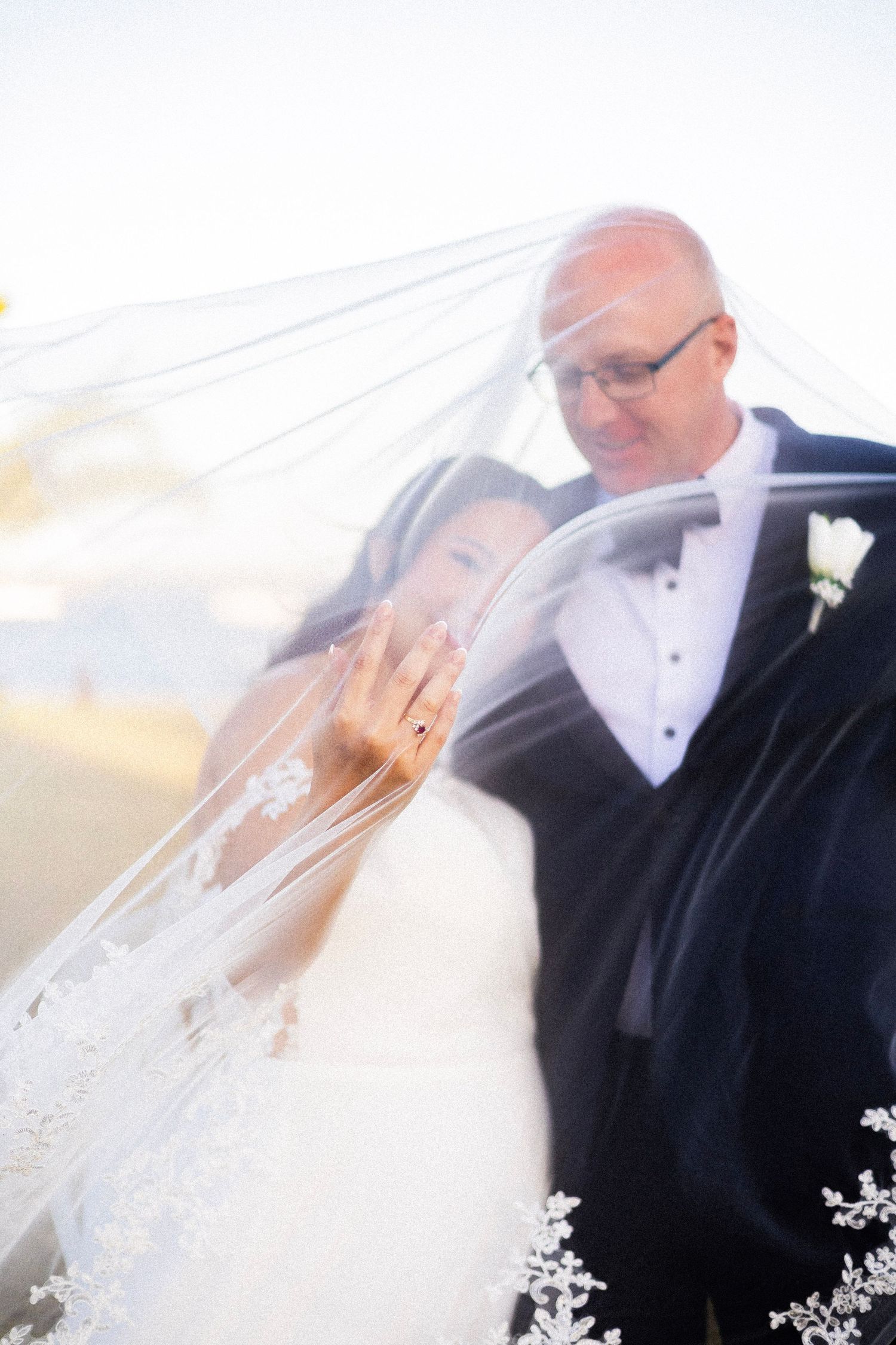 Bride shows wedding ring while couple embraces beneath flowing lace veil in dreamy soft-focus portrait.