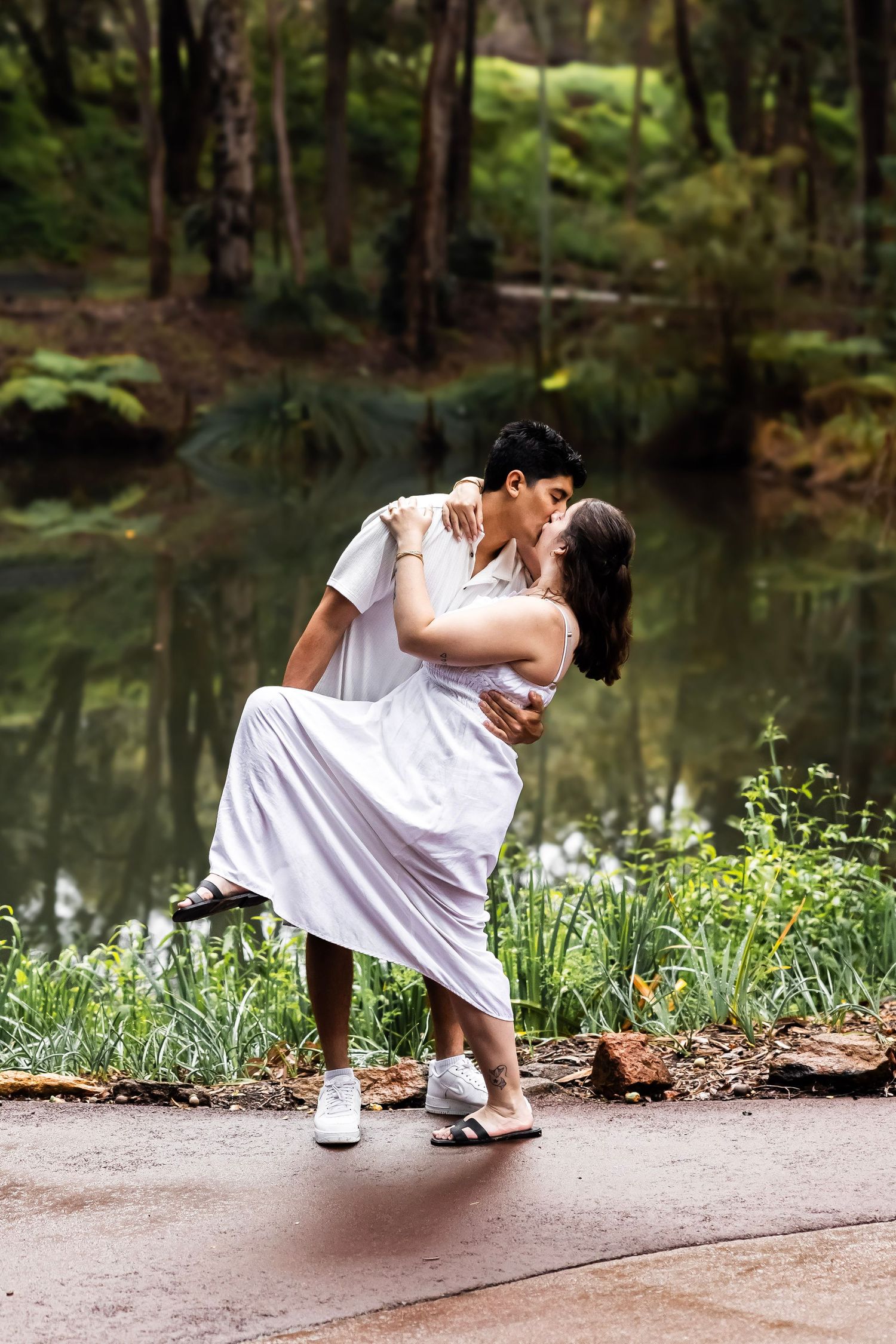 A couple shares a passionate dip kiss near a serene pond surrounded by vibrant green ferns and trees.