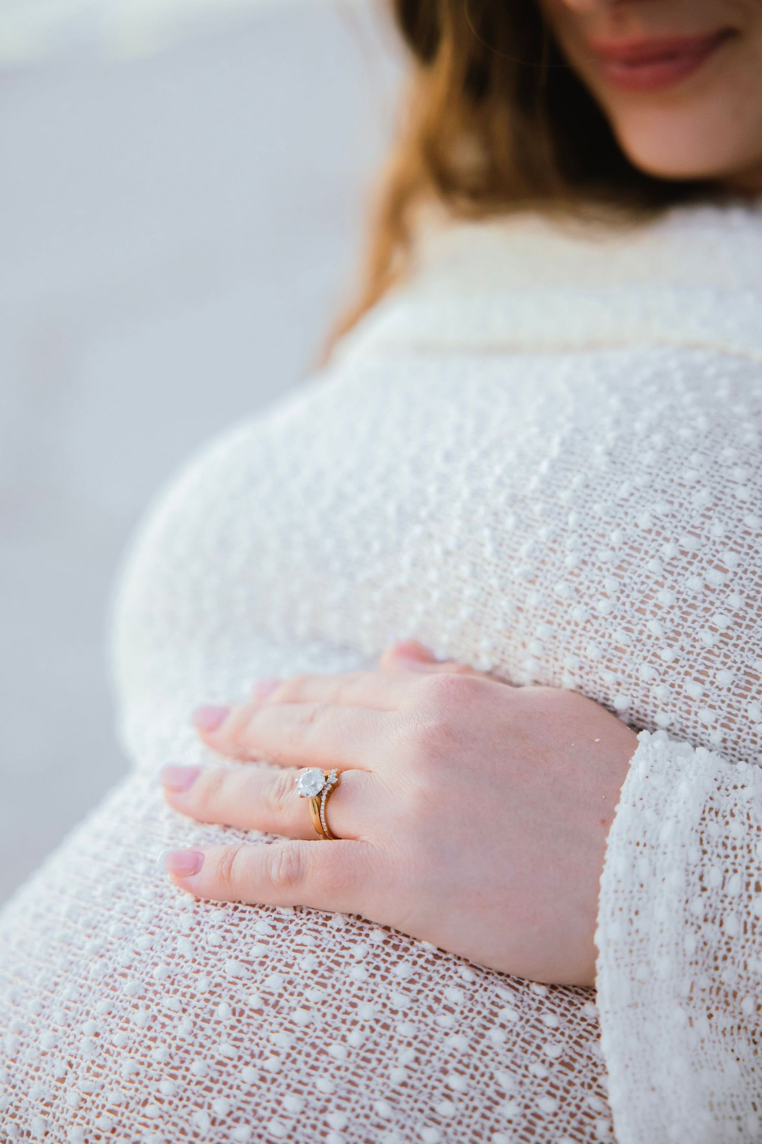 Pregnant woman in white knit sweater gently cradling baby bump, wearing gold diamond engagement ring and wedding band.