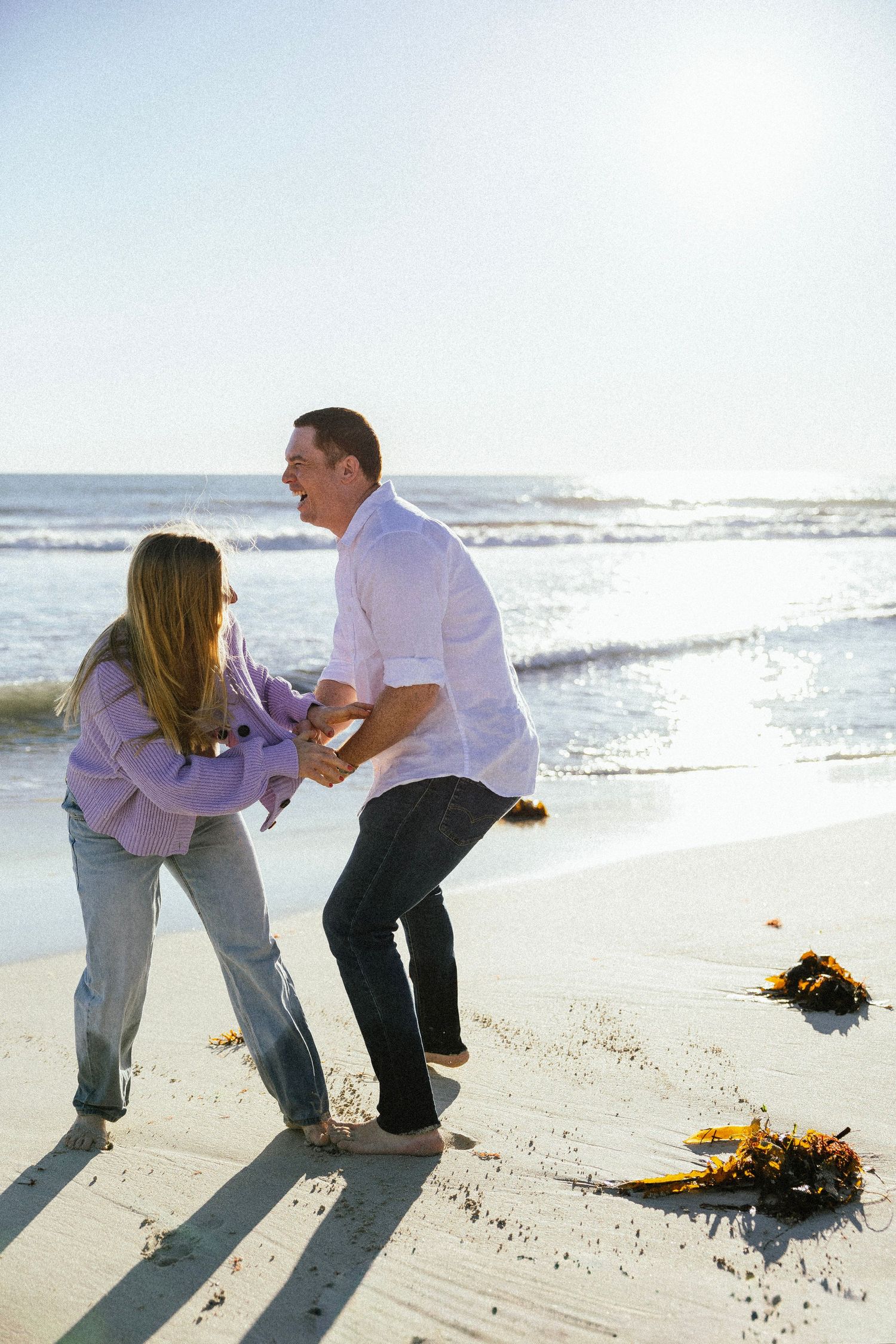 Couple laughing and dancing together barefoot on a sunny beach with ocean waves in the background.