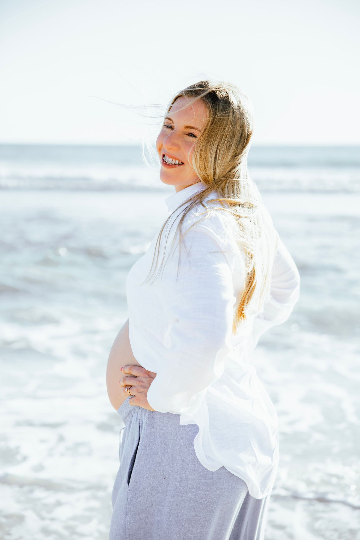 Smiling pregnant woman in white shirt holding baby bump at beach.