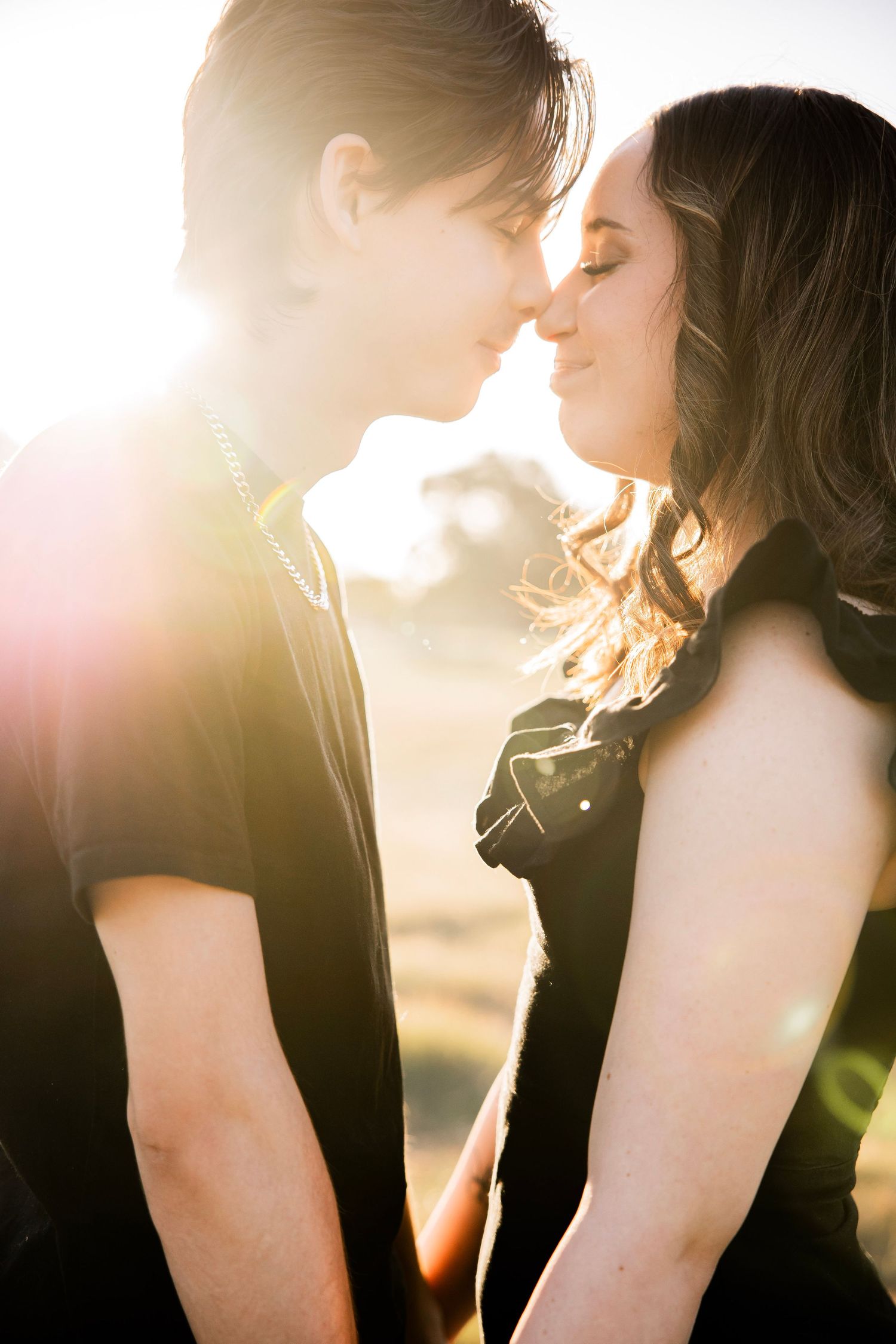 Couple leans in for a kiss backlit by golden sunset flare in an outdoor romantic portrait.