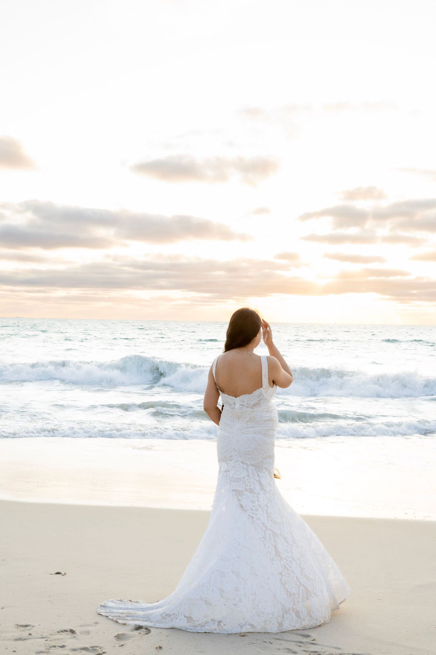 Pregnant woman in white lace mermaid gown standing with back to camera at beach sunset.
