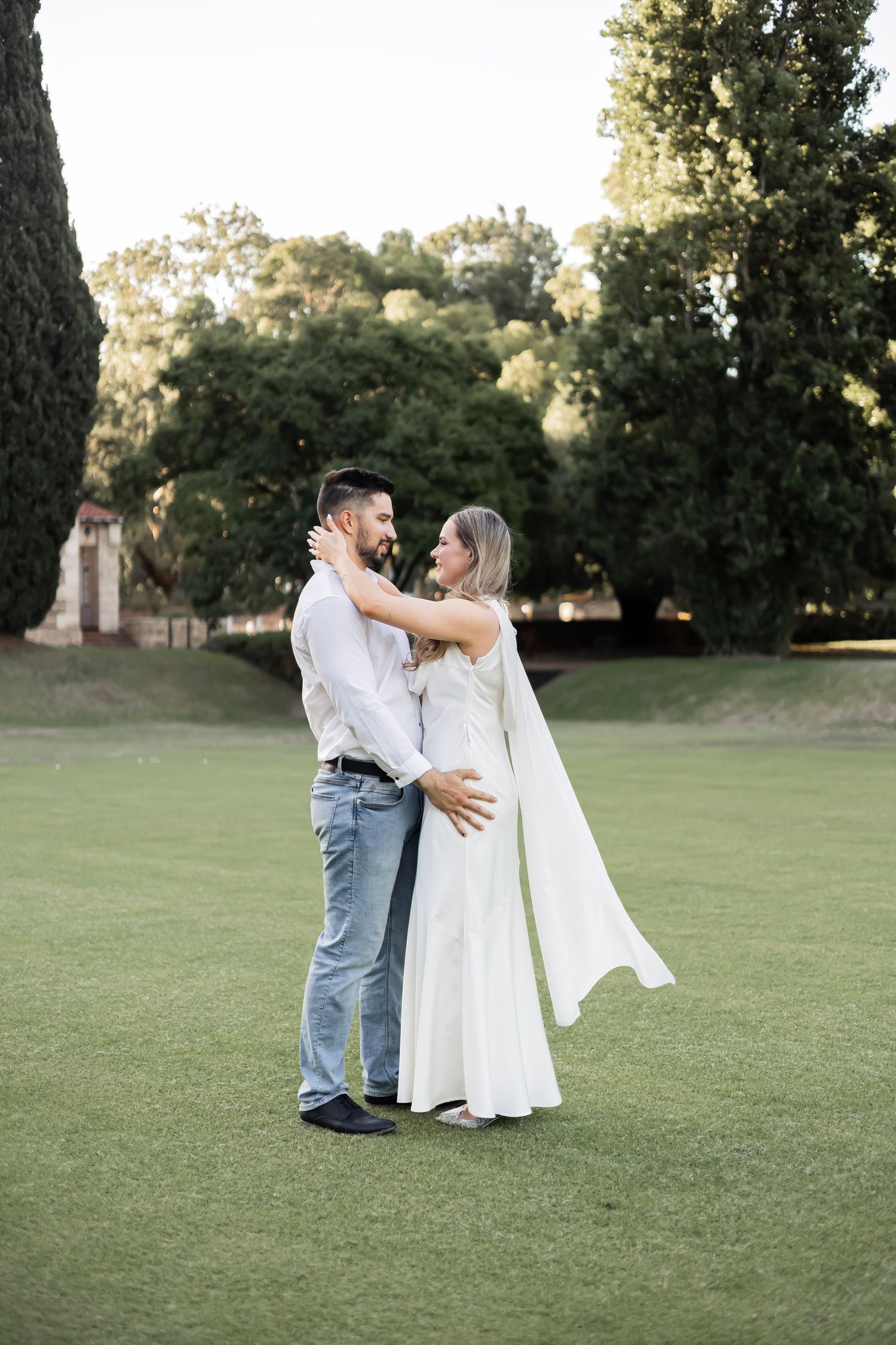 Happy couple embracing and smiling at each other on a green lawn with trees in the background.