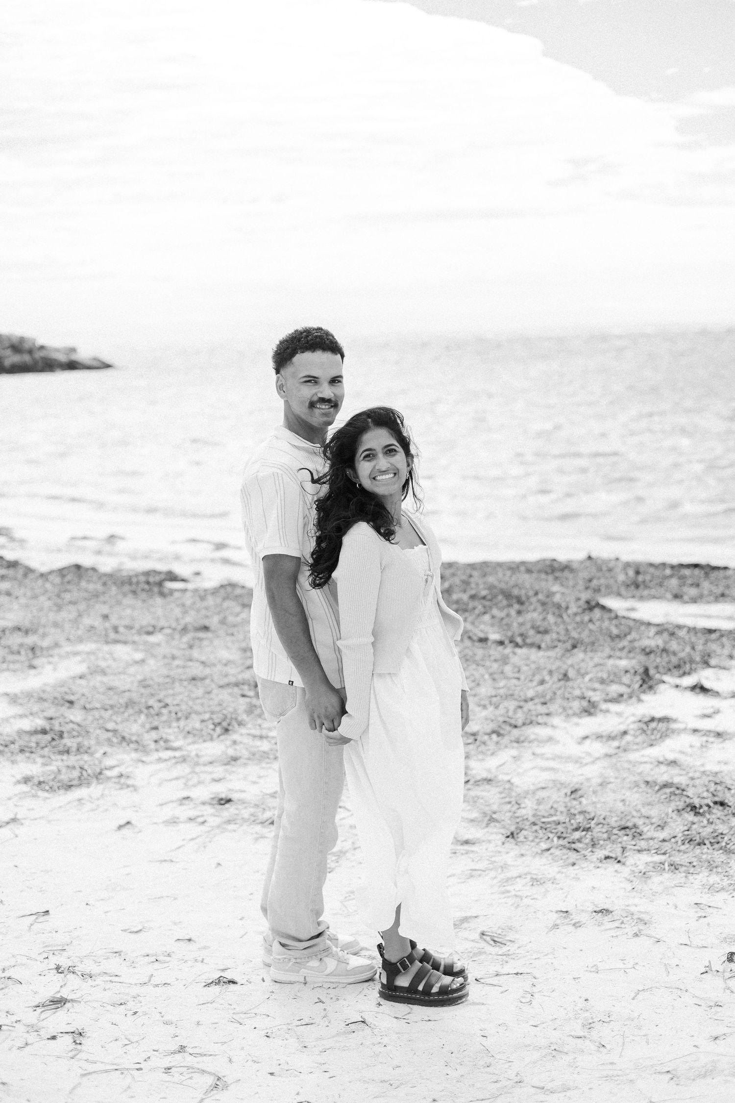 Black and white photo of a couple holding hands and smiling on a sandy beach with ocean waves behind them.