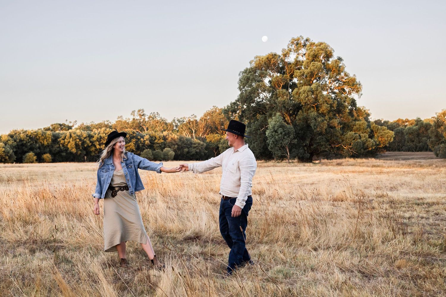 Couple holding hands and dancing in a golden field at sunset with trees and moon in background.