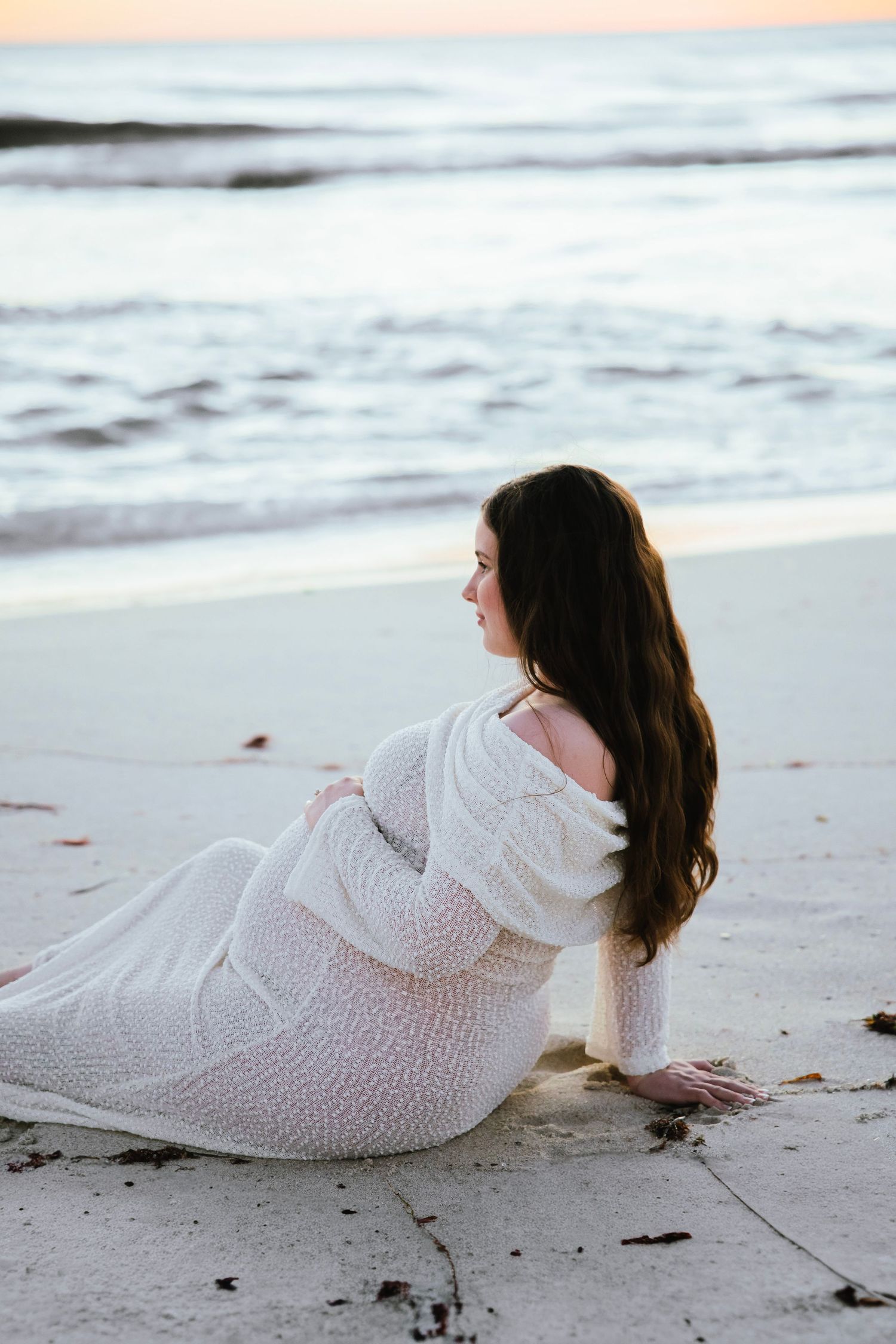Pregnant woman in white knit dress sitting on beach sand, gazing toward ocean at dusk.