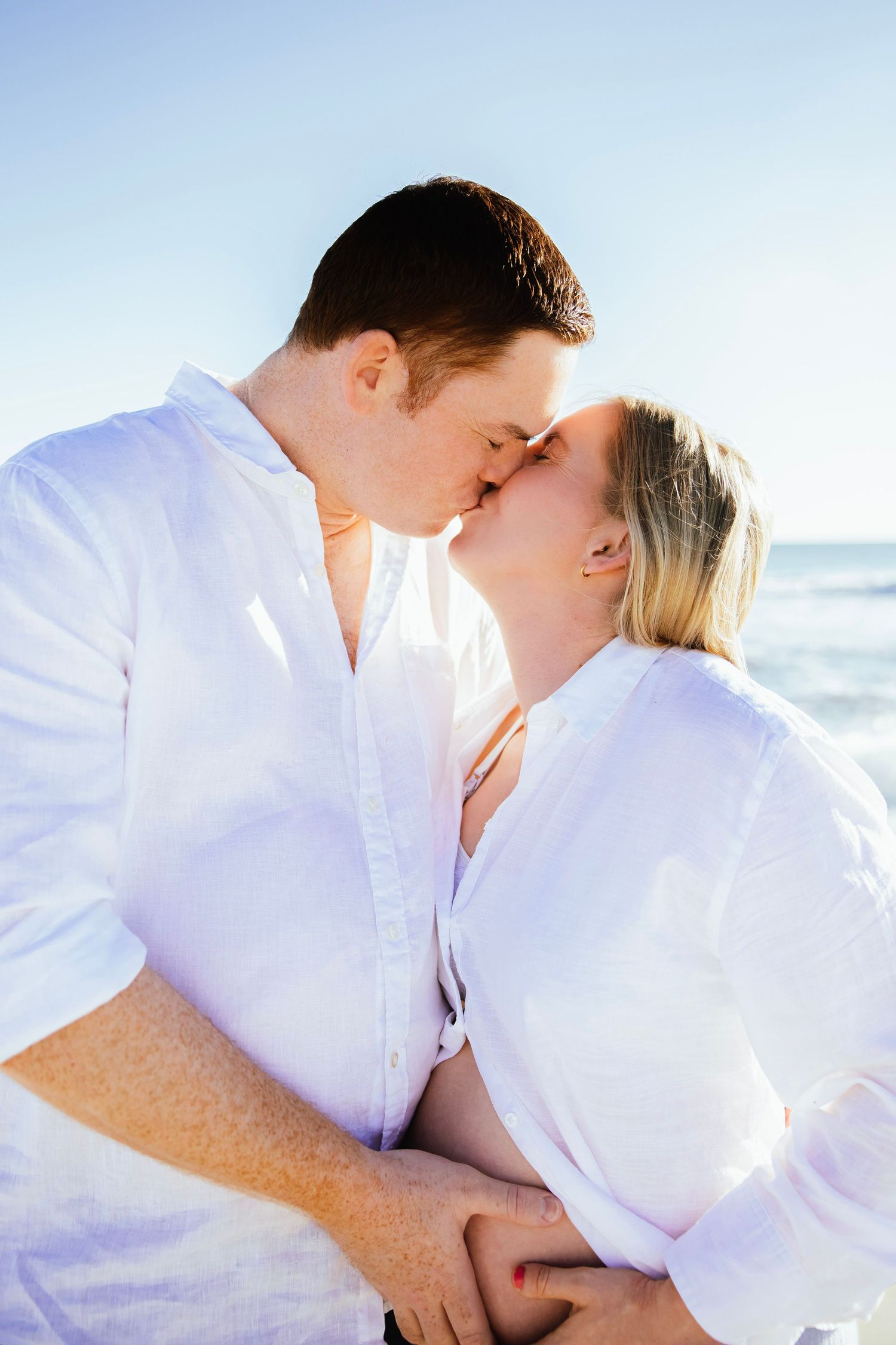 Romantic pregnant couple sharing a kiss on a bright sunny beach, both wearing white linen shirts.