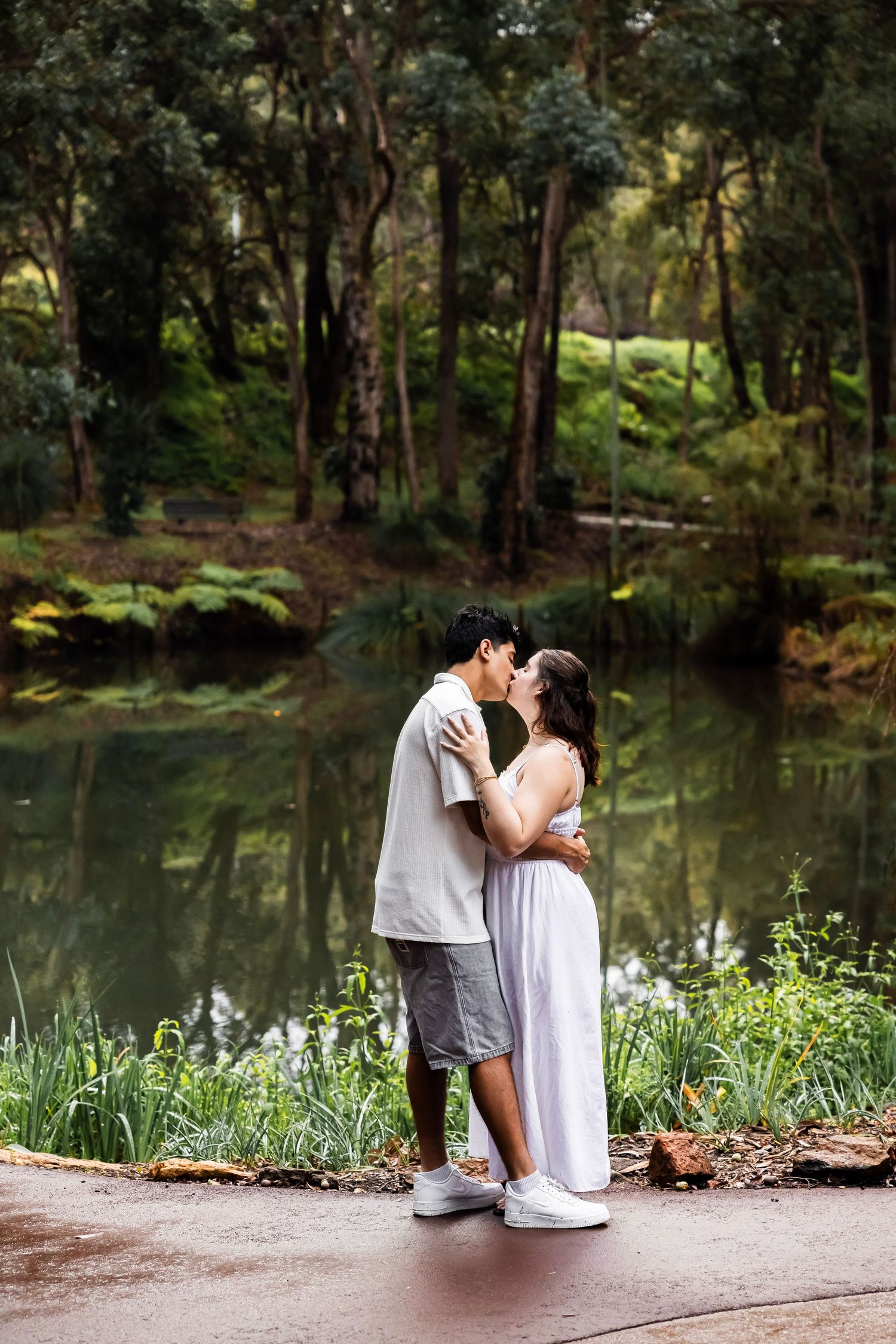 Couple kissing beside a calm reflective pond in a lush green forest park setting.
