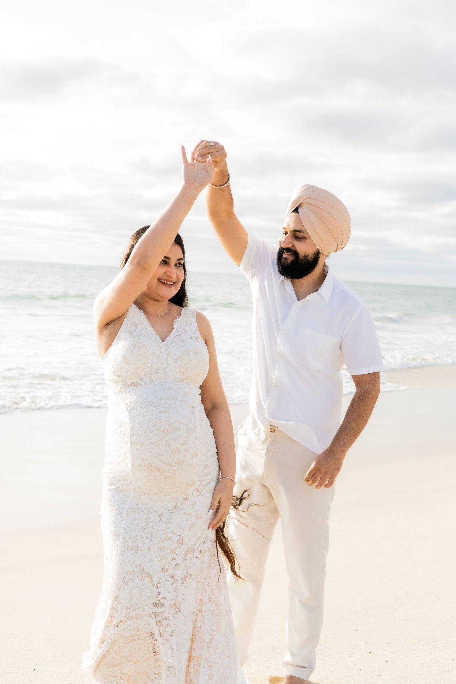 Joyful pregnant couple in white dancing on a sandy beach with ocean waves in the background.