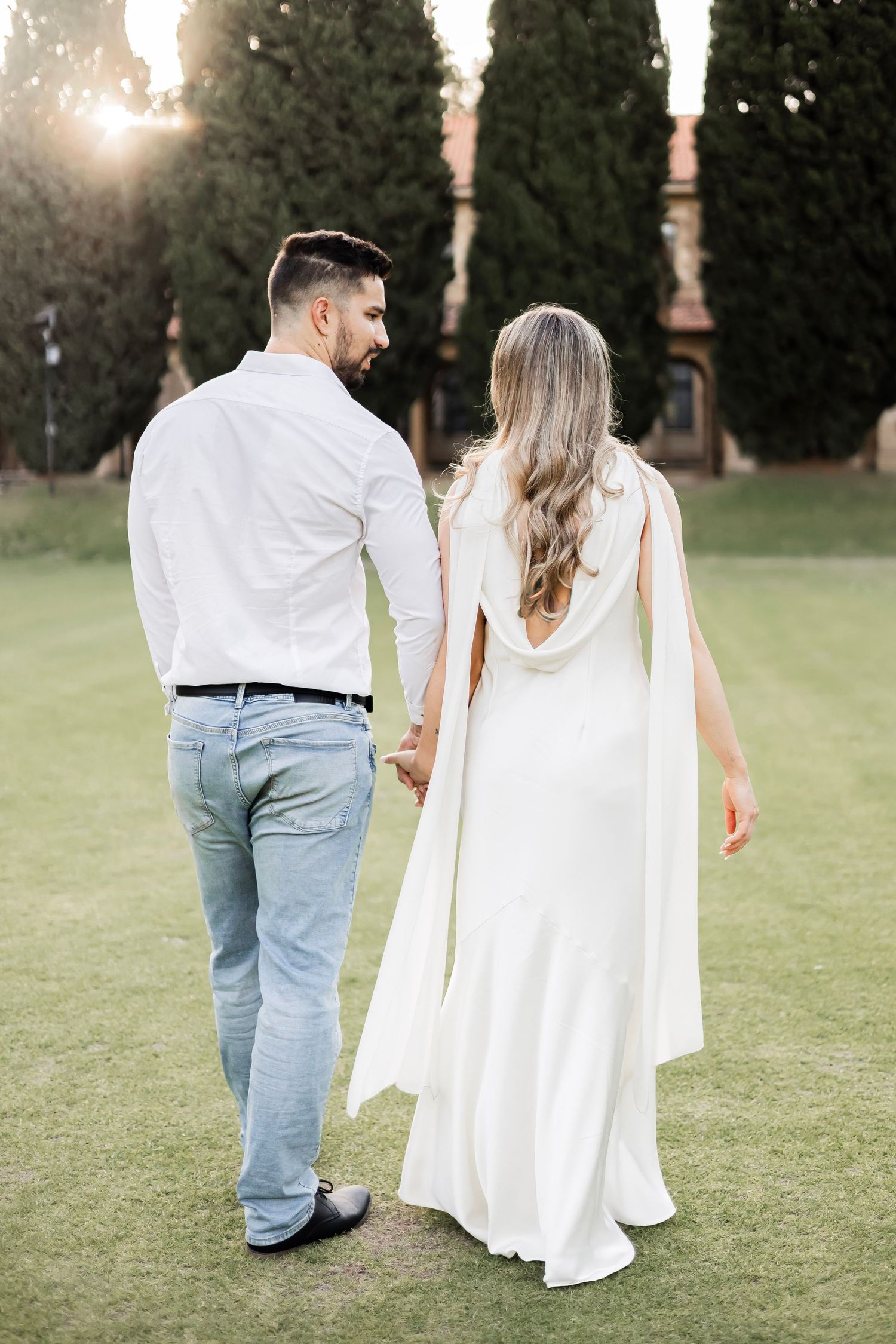 Couple holding hands walking on green lawn toward cypress trees, woman in white bridal gown, man in jeans.