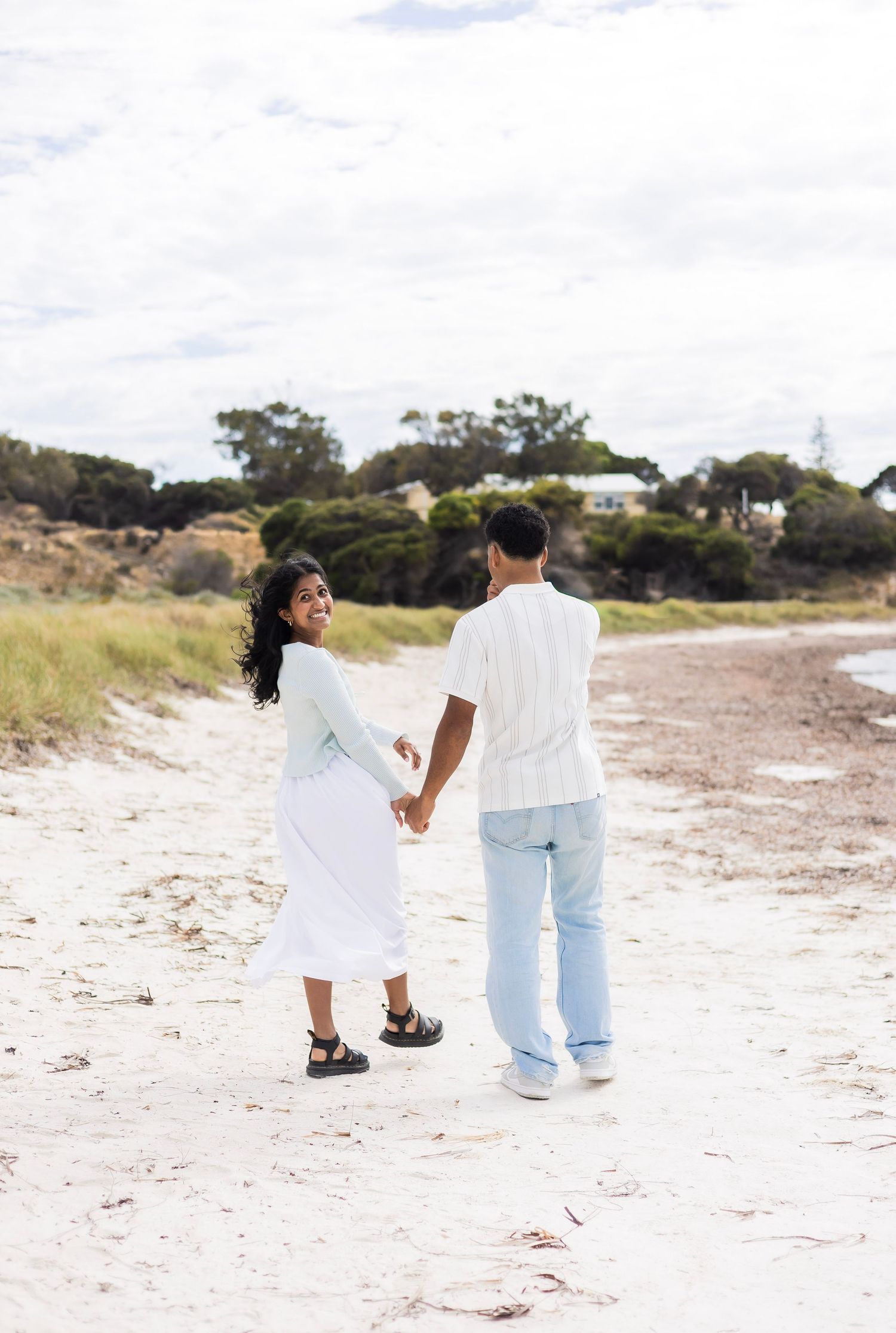 A smiling couple holds hands while walking along a sandy beach path with trees behind.