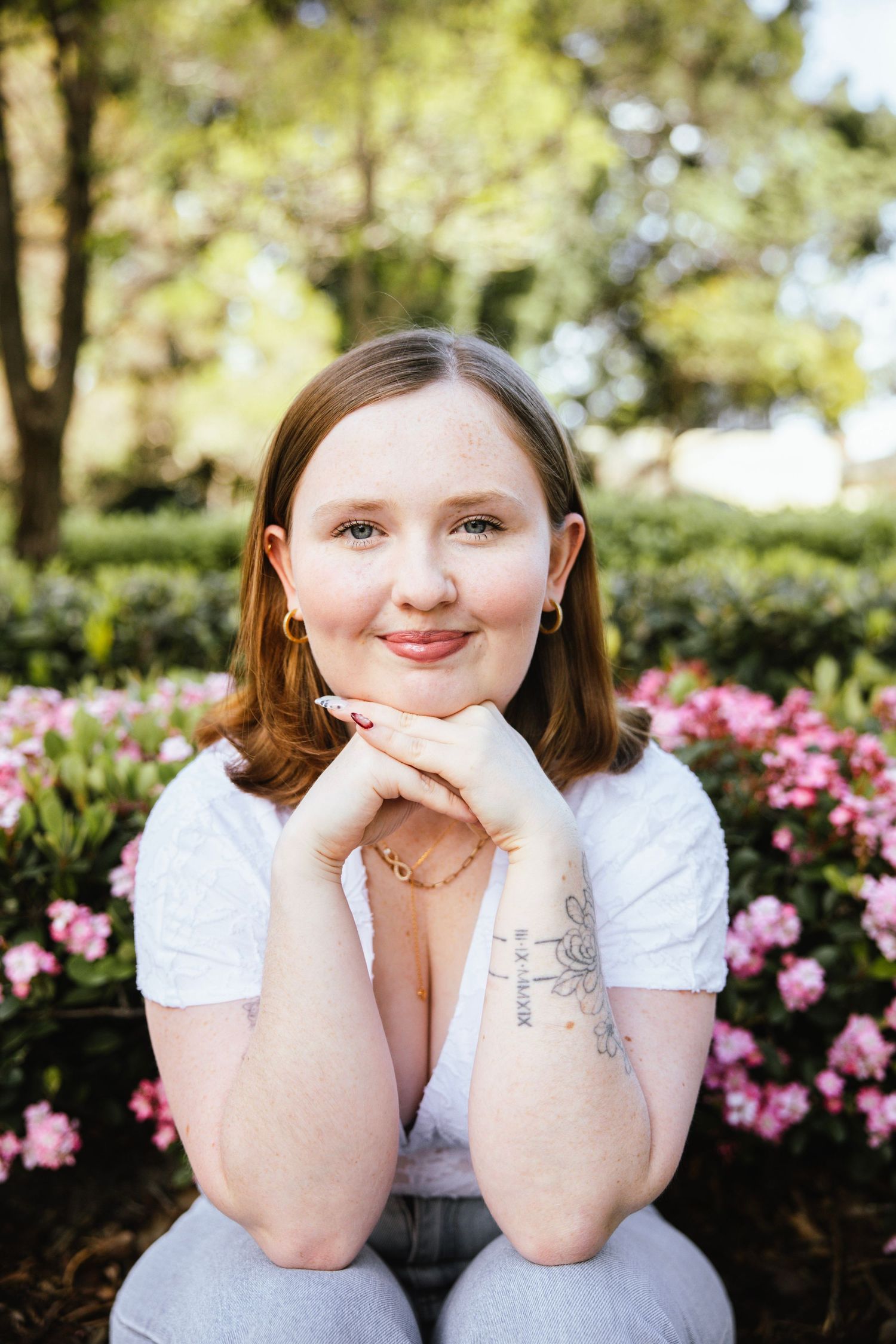Young woman with auburn hair sits among pink flowers, hands folded under chin, wearing white top with arm tattoo.