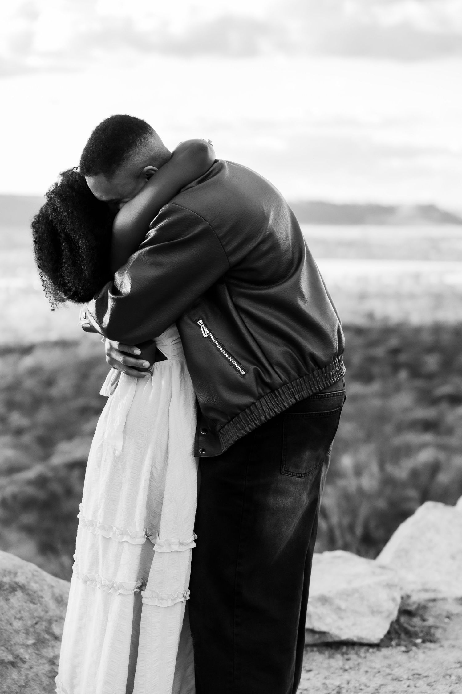 Black and white photo of a couple embracing tenderly outdoors, woman in white dress and man in leather jacket.
