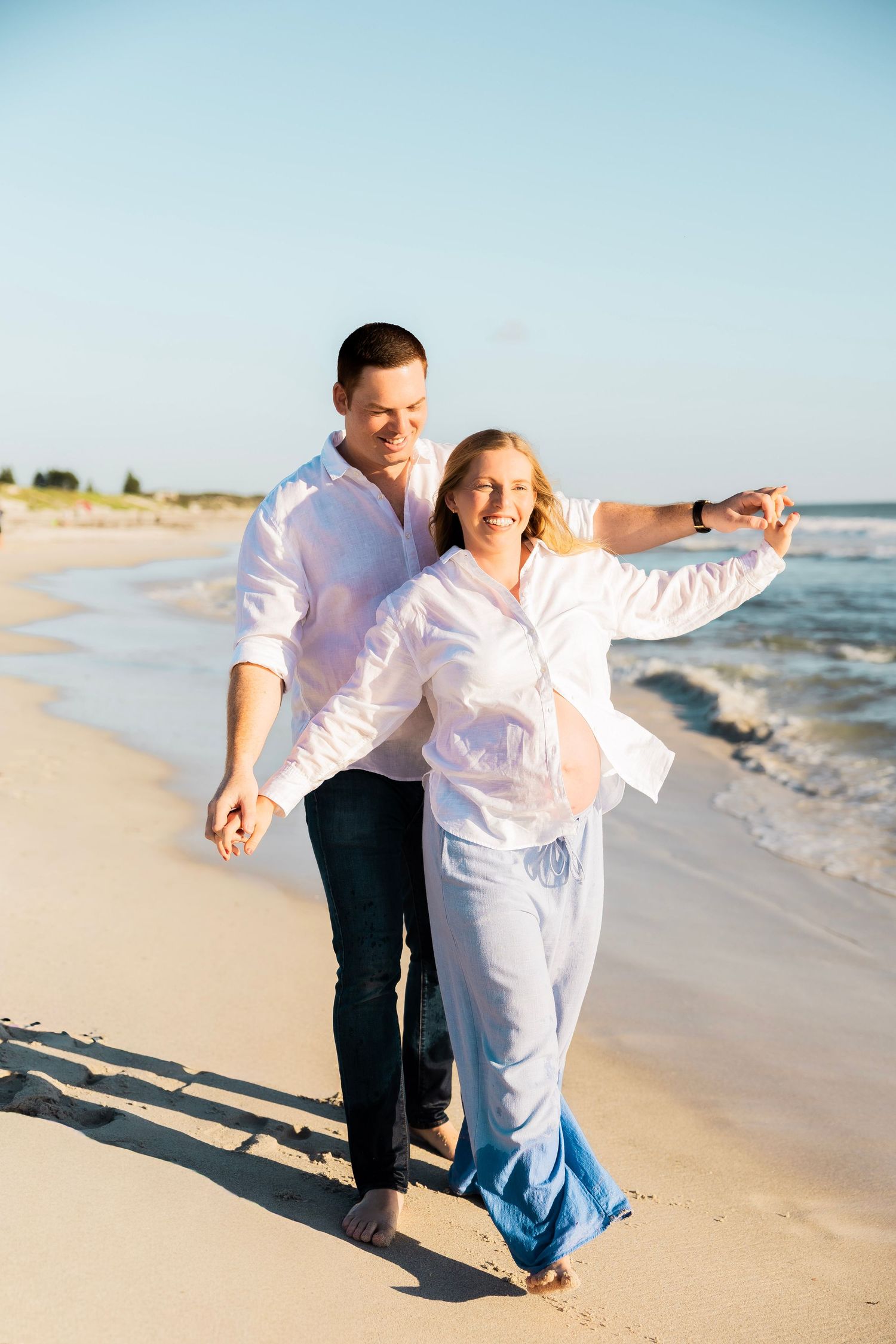 Happy couple walking hand in hand along a sunny beach, dressed in white shirts and blue pants.