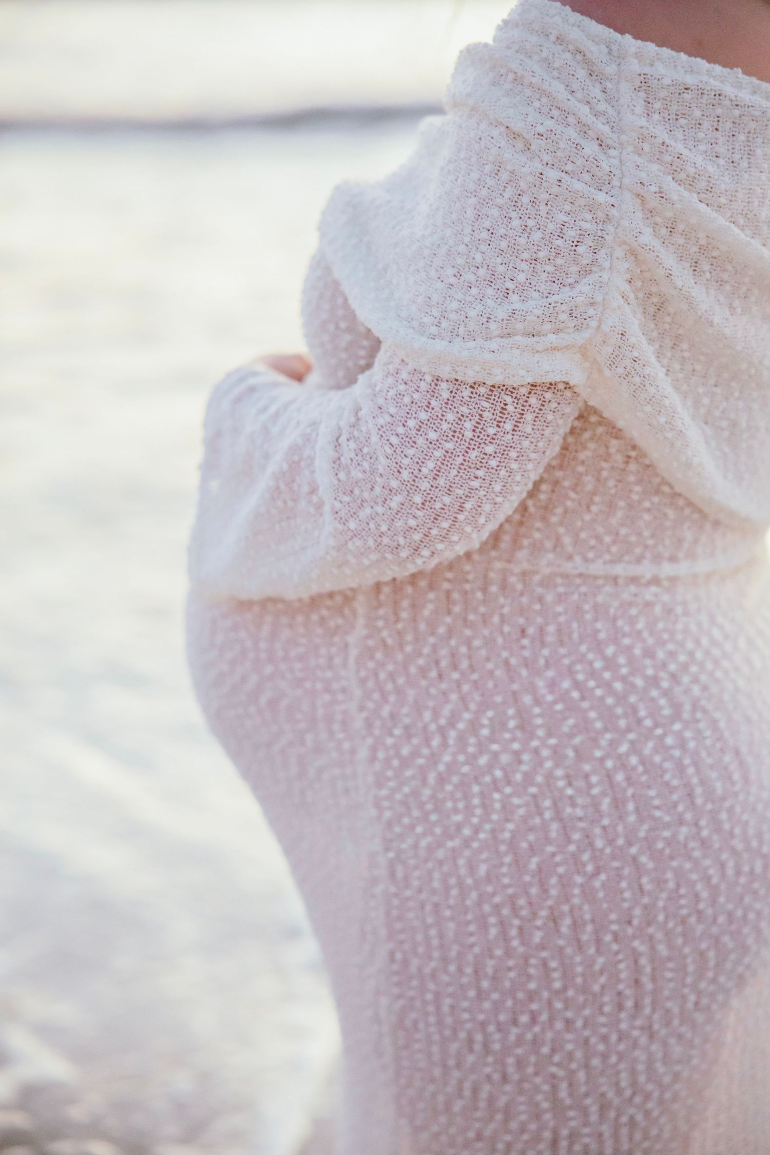 Close-up detail of white textured lace maternity dress shoulder with soft beach bokeh background.