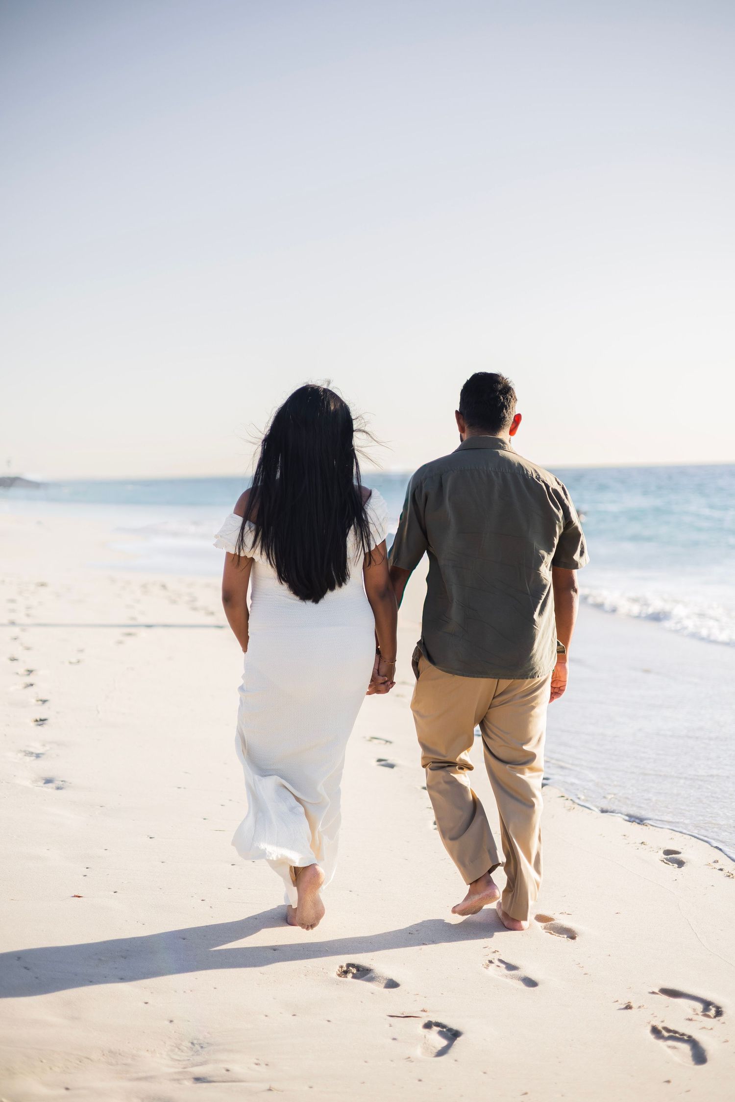 Couple holding hands walking barefoot on a sandy beach toward the ocean, leaving footprints in the sand.