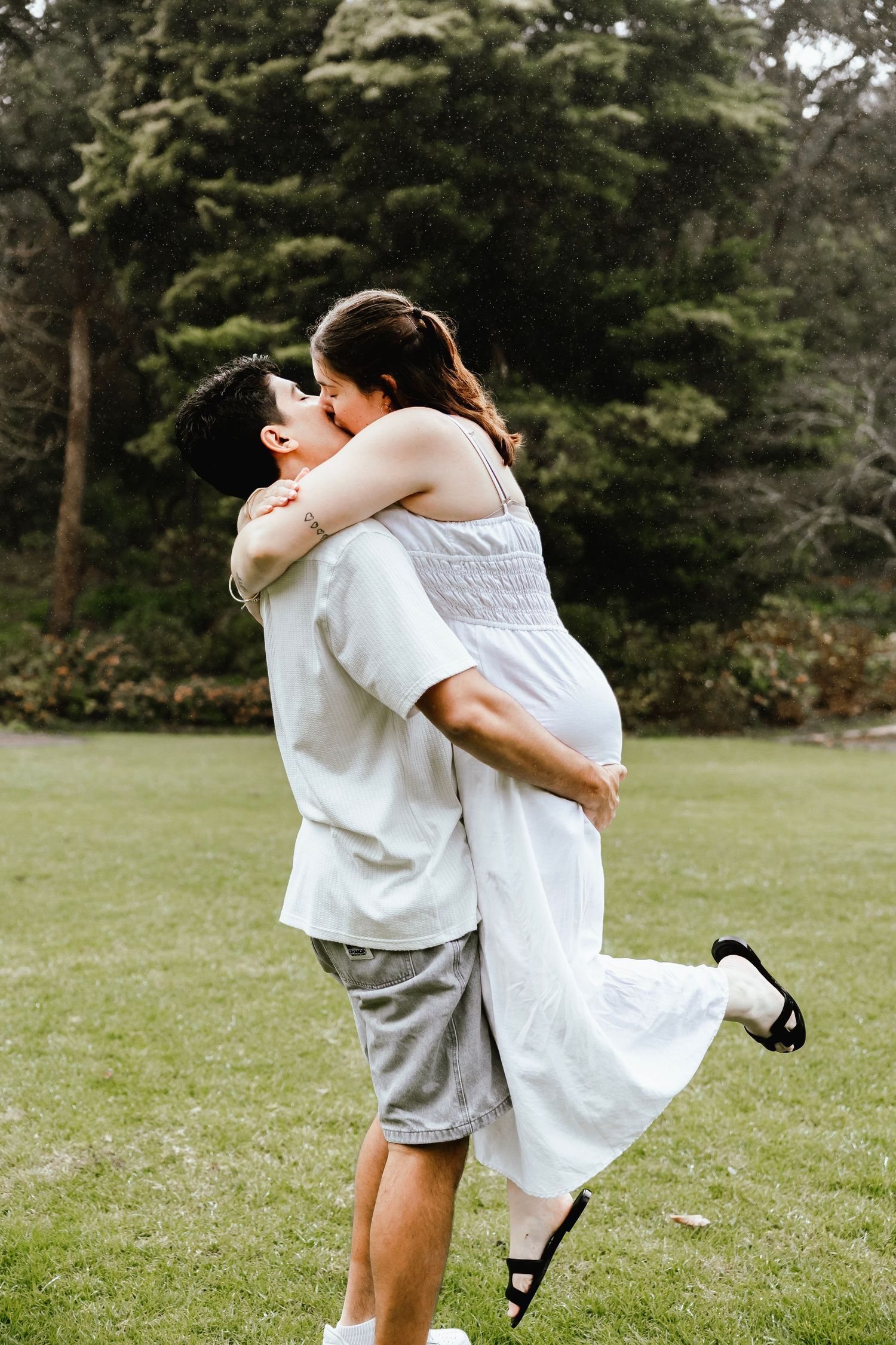 A couple shares a romantic kiss outdoors, man lifting woman in white dress, surrounded by lush green trees.