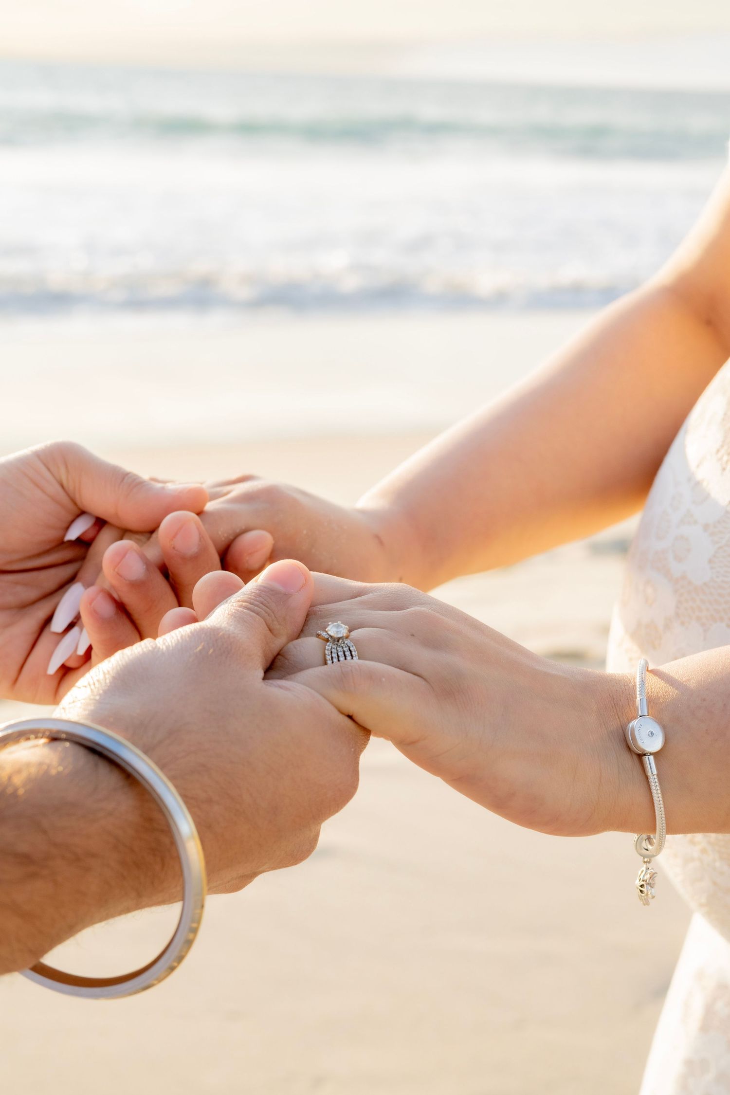 Close-up of couple holding hands on beach, woman wearing a ring and bracelet, man wearing a silver bangle.