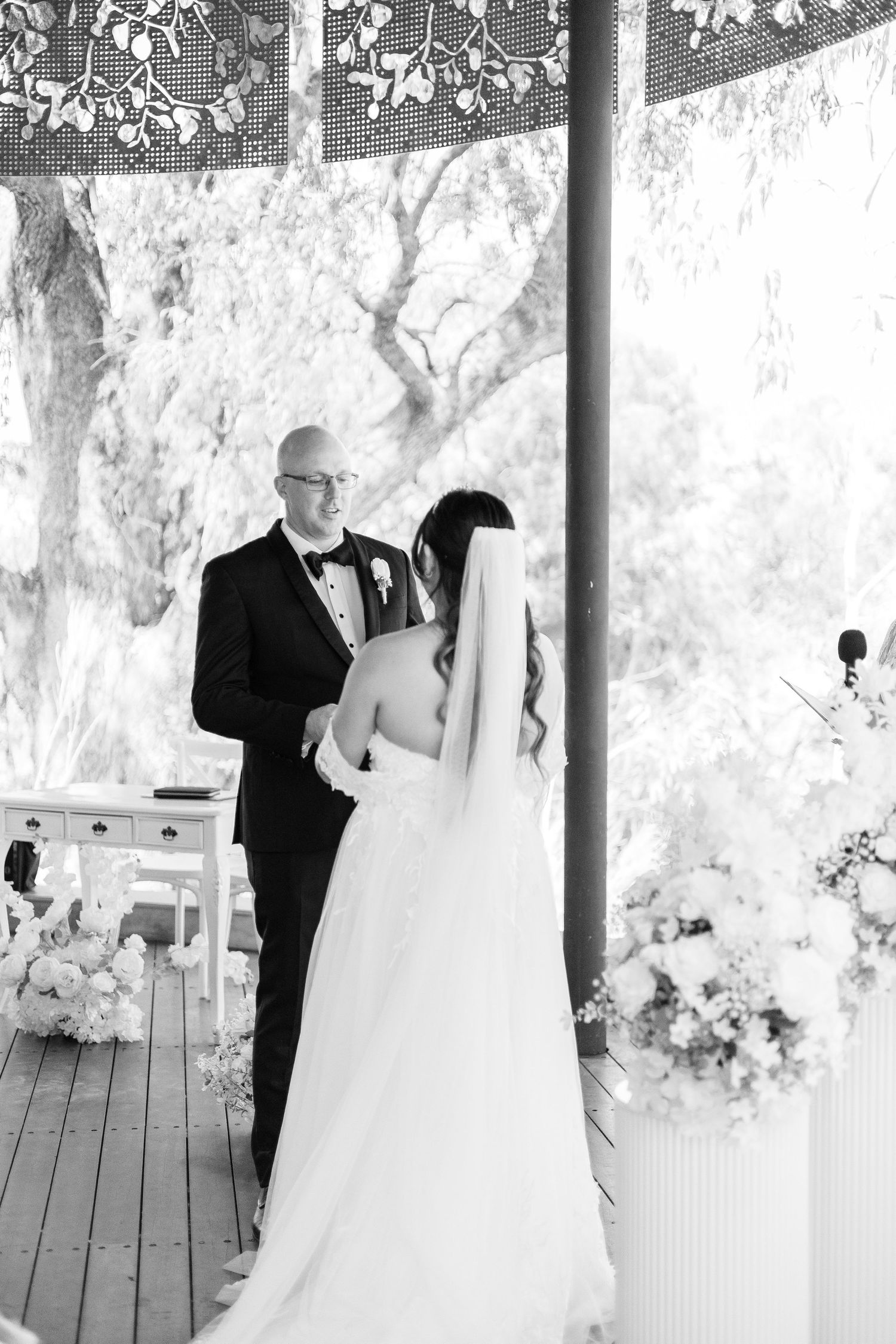 Black and white photo of bride and groom exchanging vows under a floral gazebo during an outdoor wedding ceremony.
