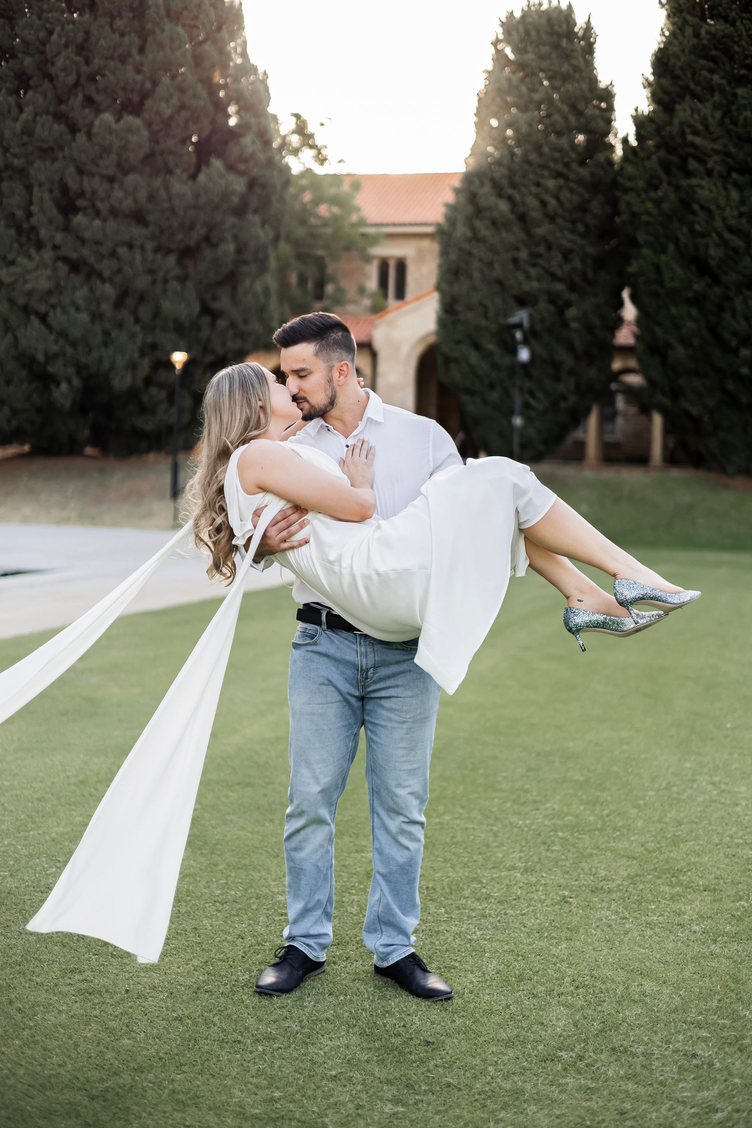 Groom carrying bride in white dress and veil, sharing a kiss outdoors on green lawn with trees and building behind.