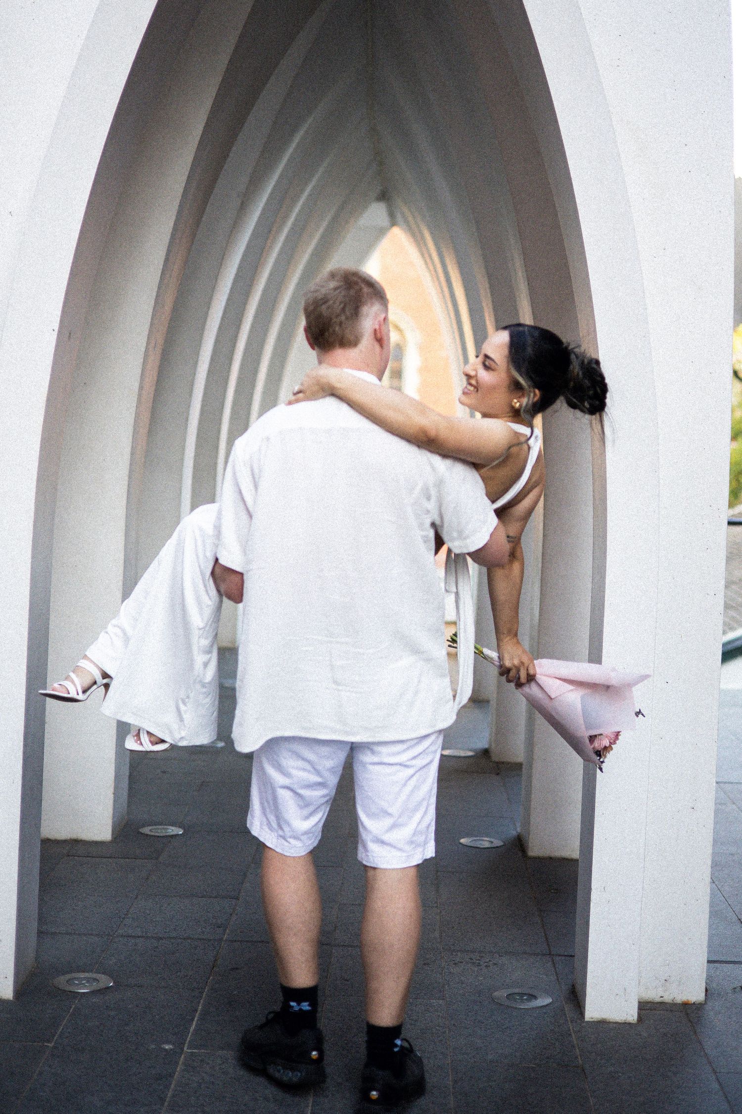 Man in white carrying woman in white dress under arched walkway, romantic couple photo.