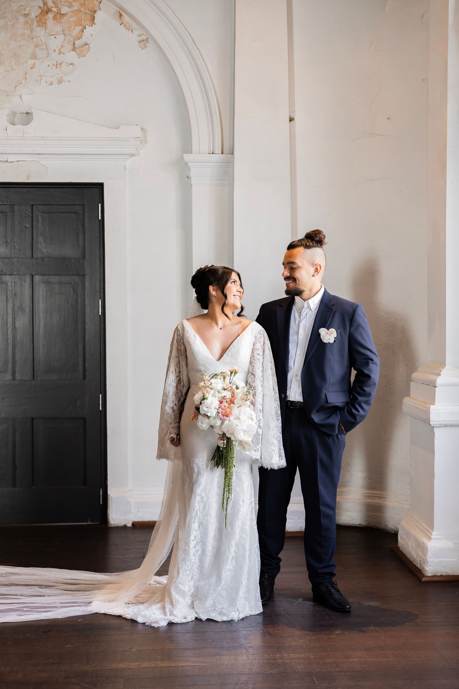 Bride in lace gown and groom in navy suit share a tender glance inside elegant white-walled venue.