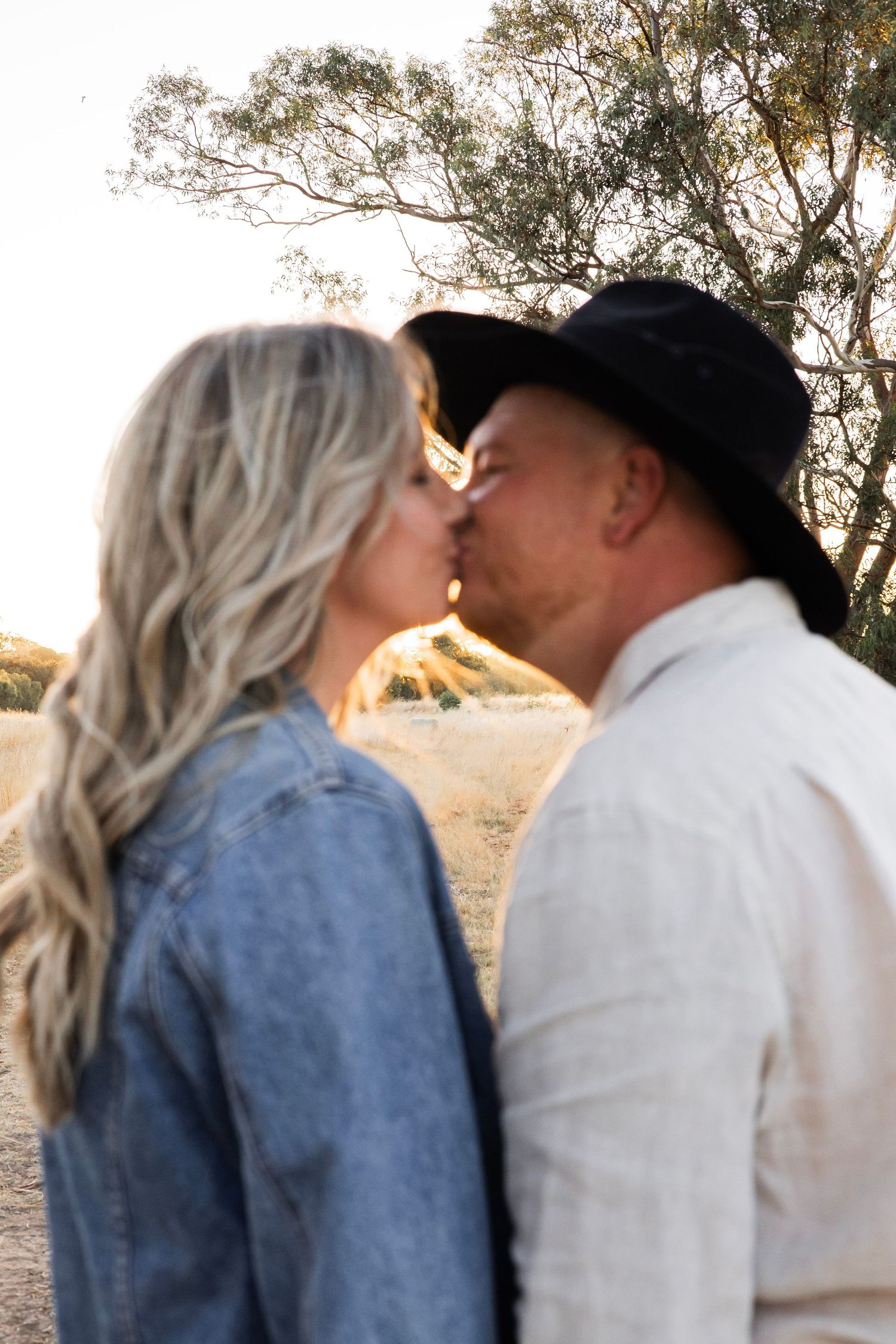 Close-up of couple sharing a kiss outdoors, man wearing black cowboy hat, warm natural lighting.