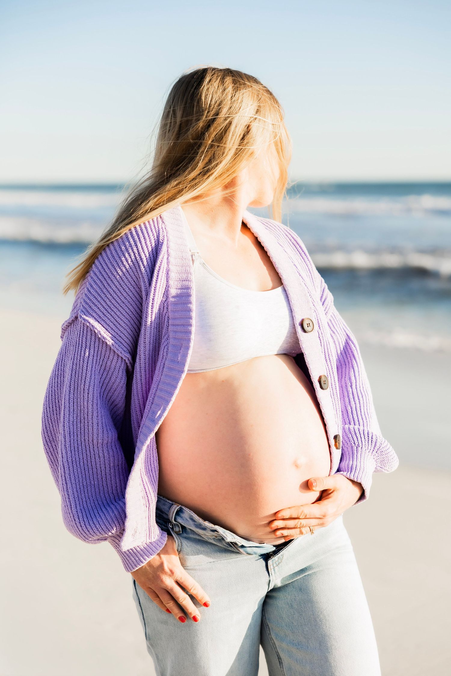 Pregnant woman in purple cardigan and jeans cradling bare baby bump on sunny beach with ocean behind.