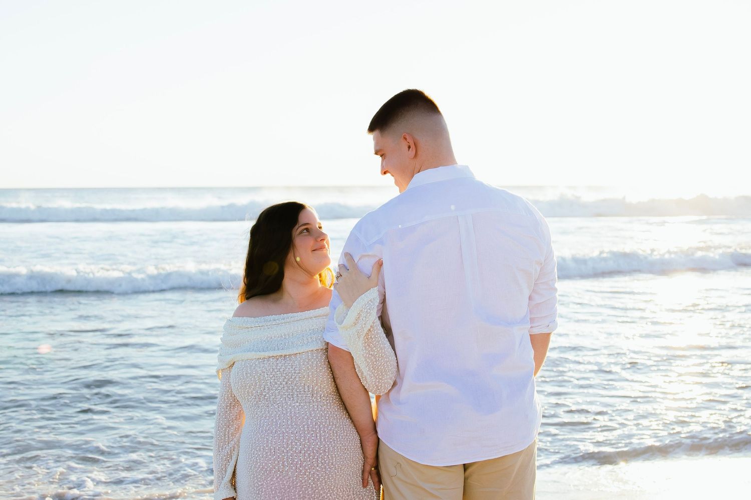 Couple standing together on a sunny beach, gazing at each other with ocean waves in the background.