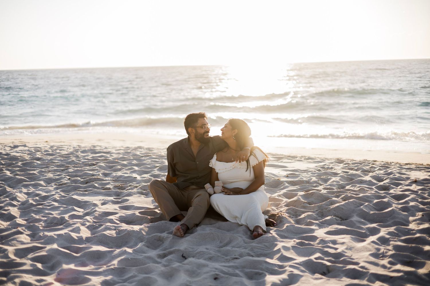 Couple sitting together on sandy beach at sunset, sharing a romantic moment by the ocean waves.